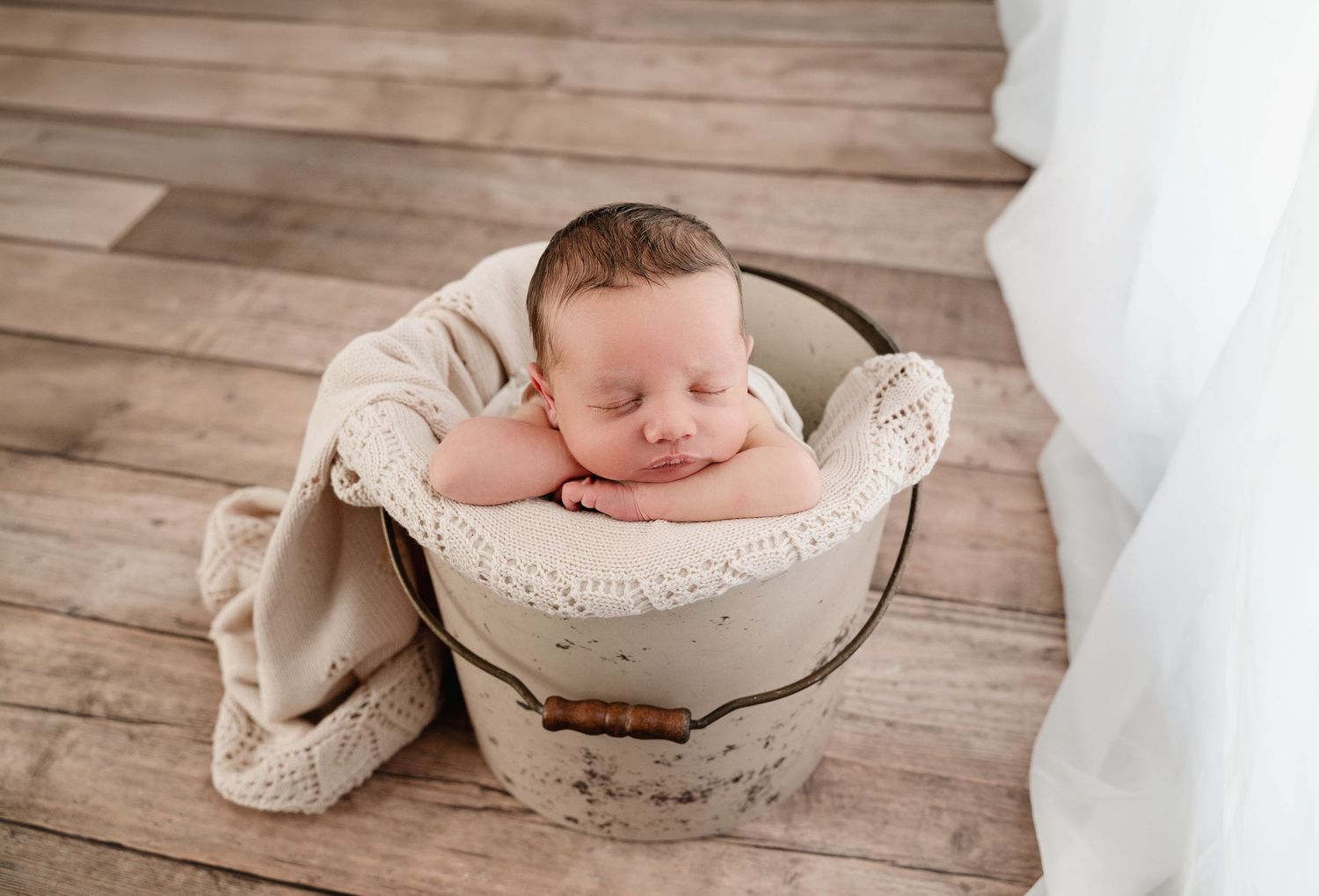 A sleeping baby nestled in a rustic white bucket on wooden flooring with a cozy beige blanket.