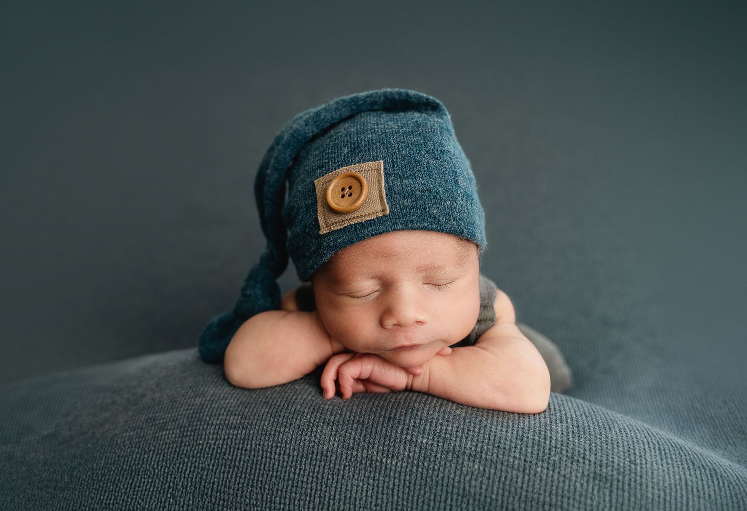 Peaceful sleeping newborn wearing blue knit beanie rests on gray blanket in professional portrait setting.