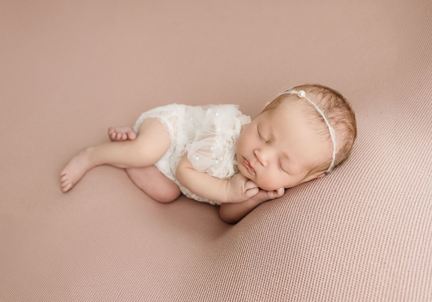 A peaceful newborn baby wearing white lace outfit sleeps on soft pink blanket with delicate headband.