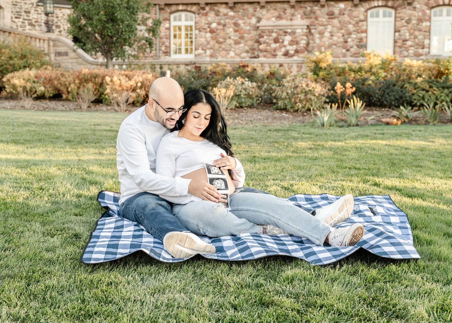 A couple relaxes together on a blue checkered blanket in a sunny grassy field during a picnic.