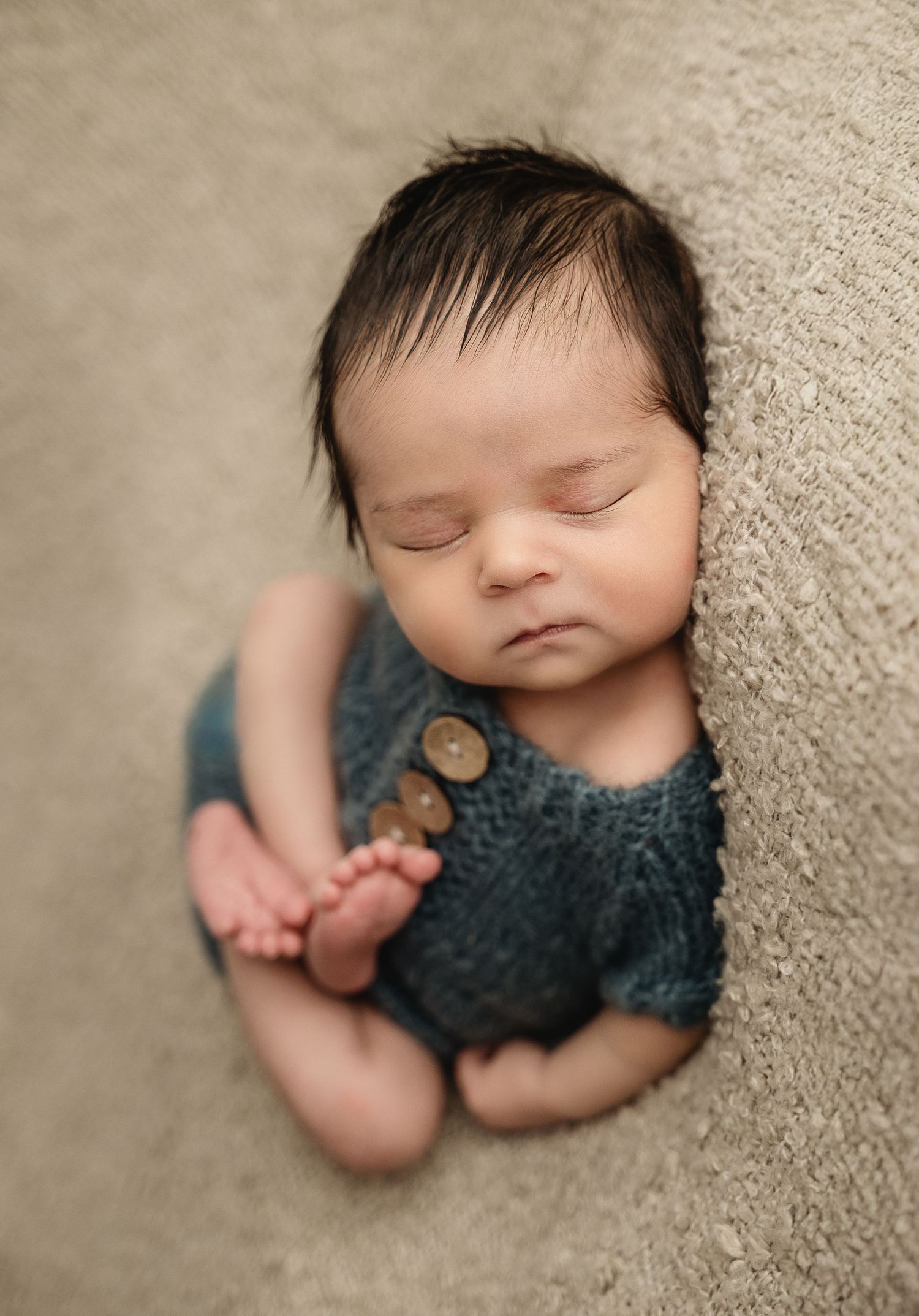 Peaceful sleeping newborn wearing knit teal romper on neutral backdrop.
