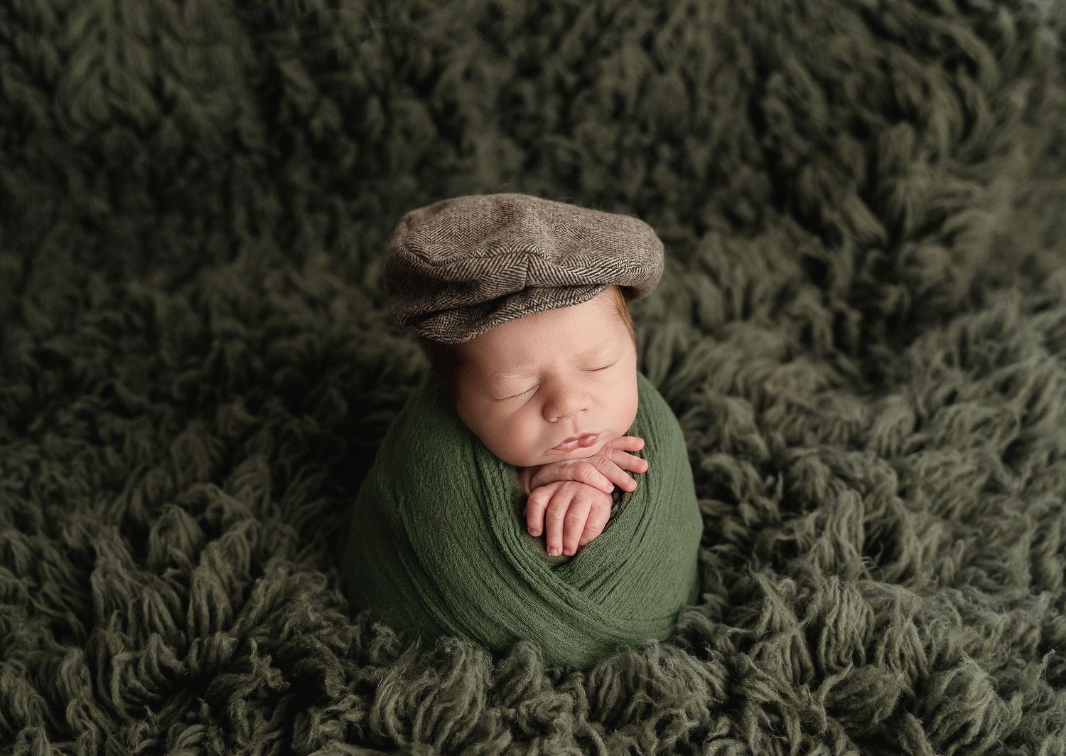 Newborn wearing flat cap and green wrap sleeping on textured green background.