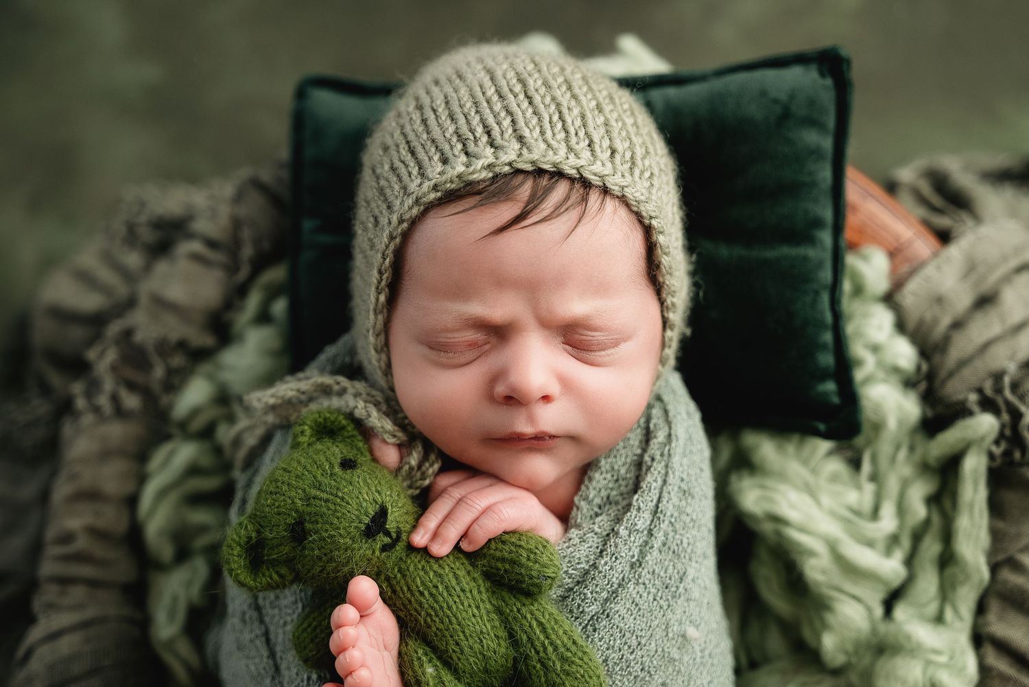 A sleeping newborn wearing a soft knit beanie and wrapped in cozy green swaddle blankets among textured fabrics.