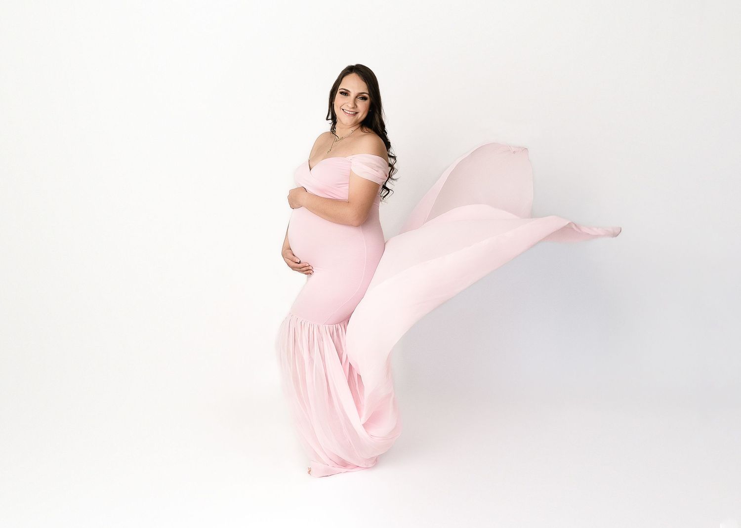 Pregnant woman in flowing pink gown poses elegantly against white background as fabric dramatically billows behind her.