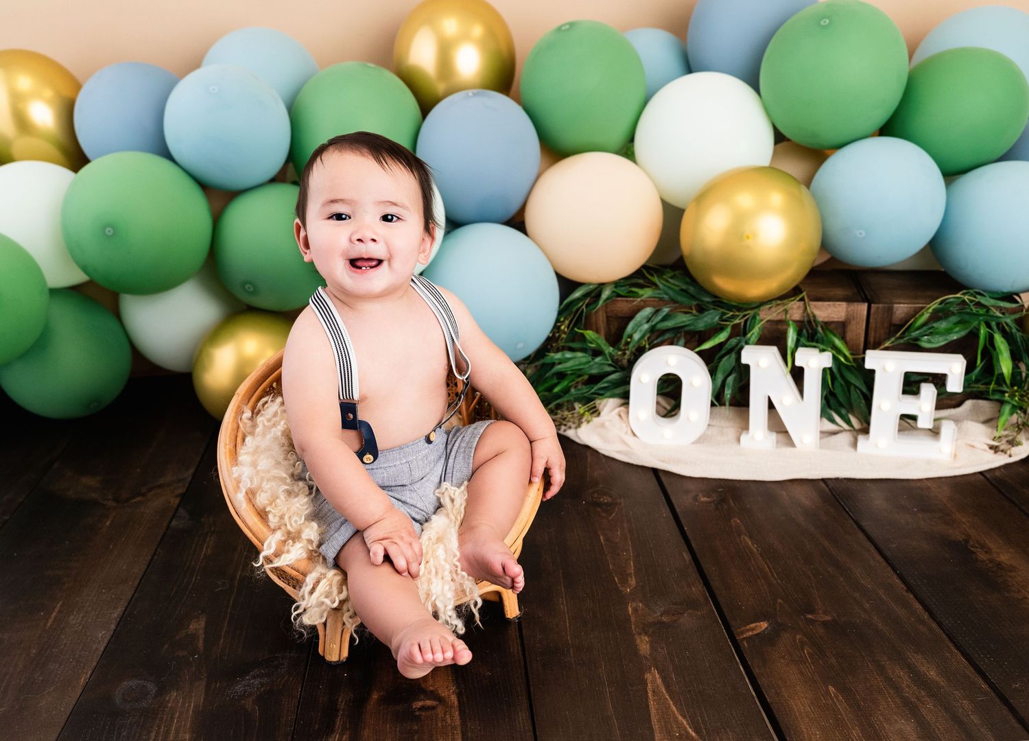Baby boy smiling in a basket at a first birthday cake smash with green, blue, and gold balloons and ONE sign.
