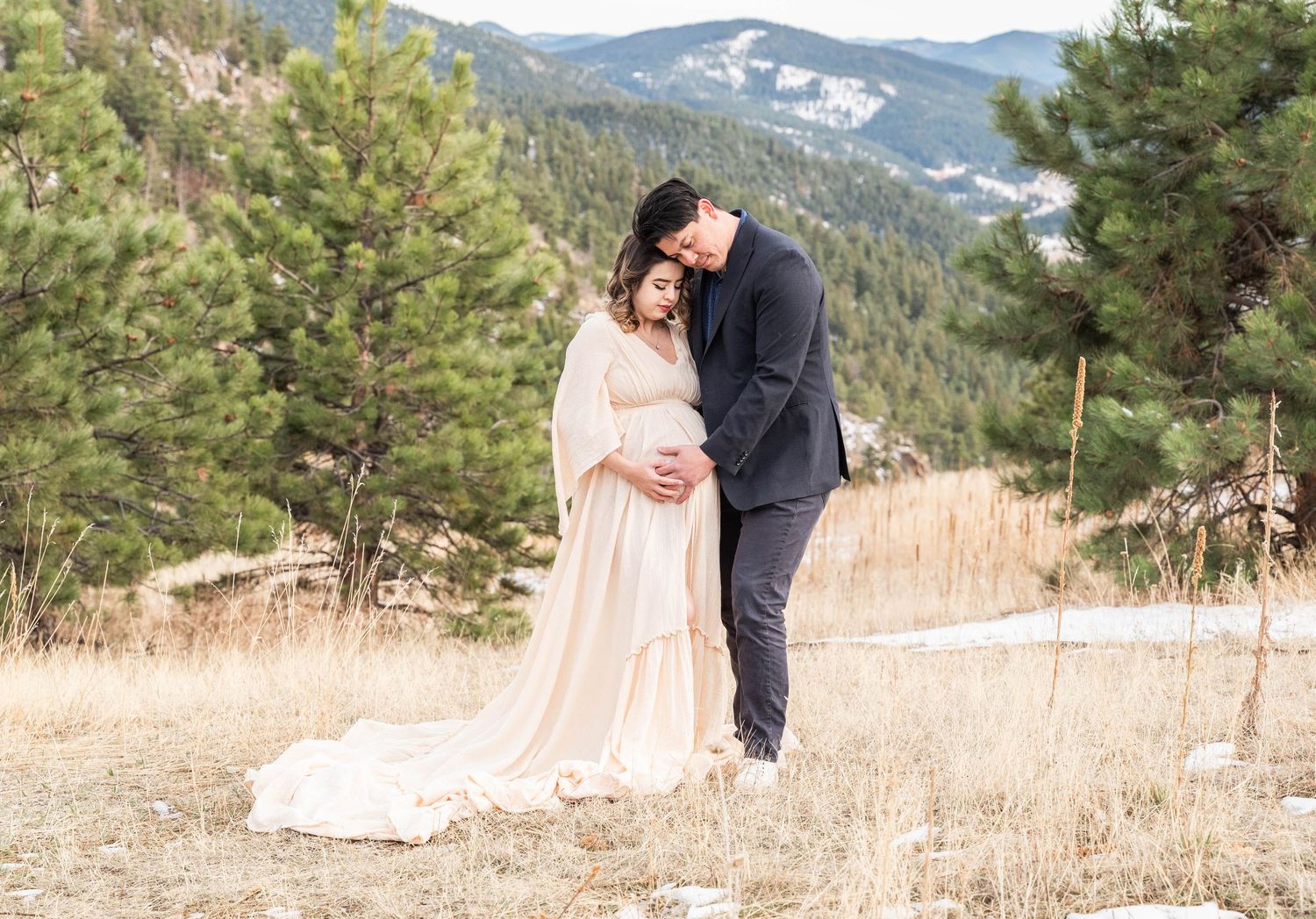 A couple in formal attire share a tender moment in a mountain meadow surrounded by evergreen trees and rolling hills.