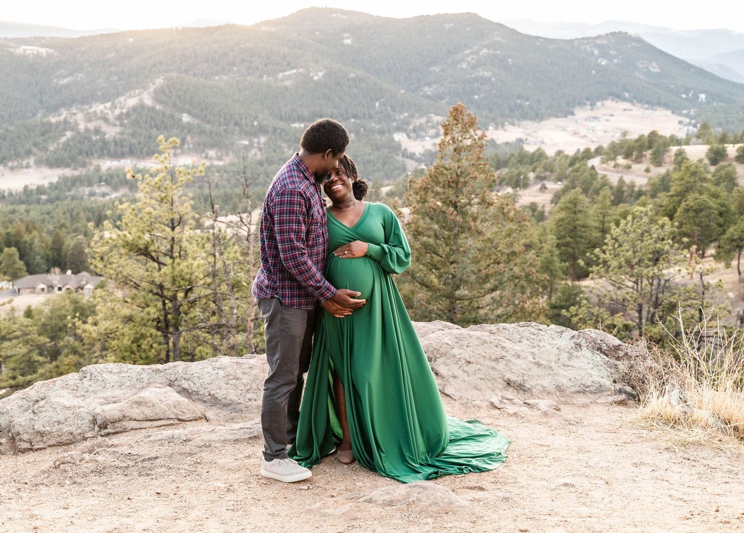 A couple stands together on a rocky mountain overlook with sweeping mountain views in the background.