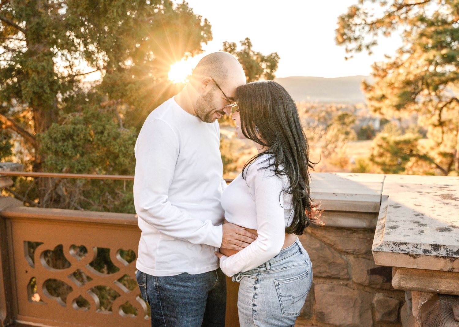 Romantic couple embraces during golden hour sunset at scenic overlook with stone wall and mountain views.