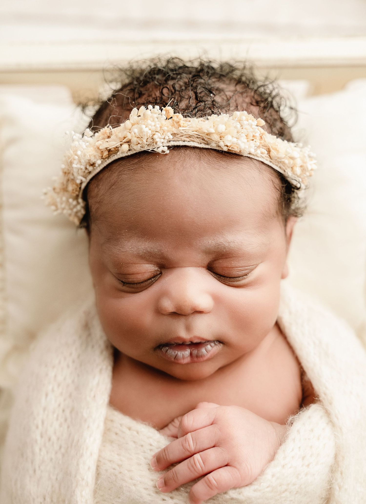 Sleeping newborn baby girl wearing a delicate floral headband, wrapped in a cream knit blanket.