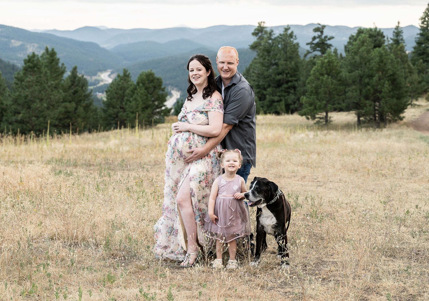 People wearing floral dresses stand in a sunny meadow with mountains and trees in the background.