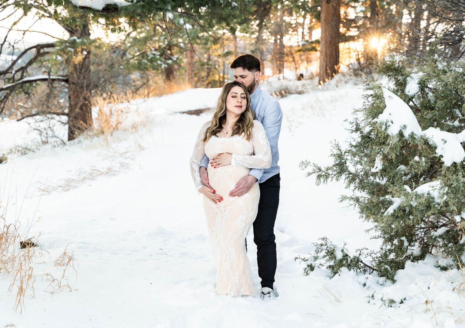 A couple poses together in a snowy winter forest setting during golden hour sunset.
