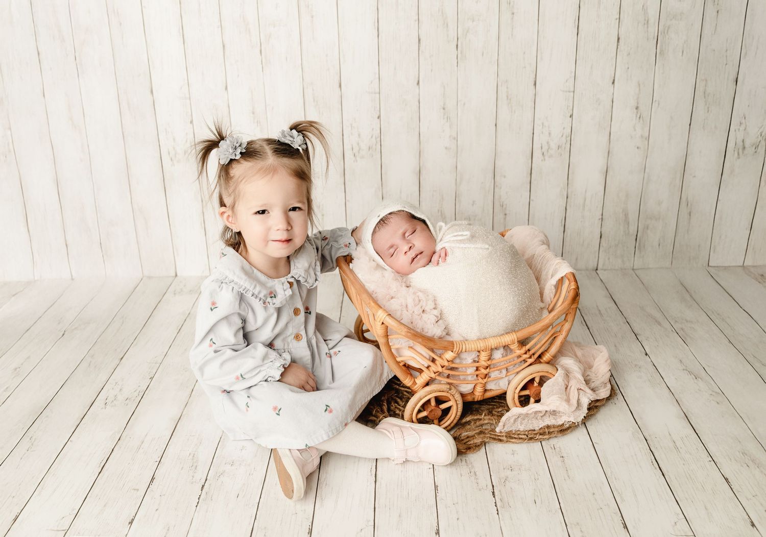 Toddler in gray dress sits beside woven basket with sleeping newborn on white wooden floor backdrop.