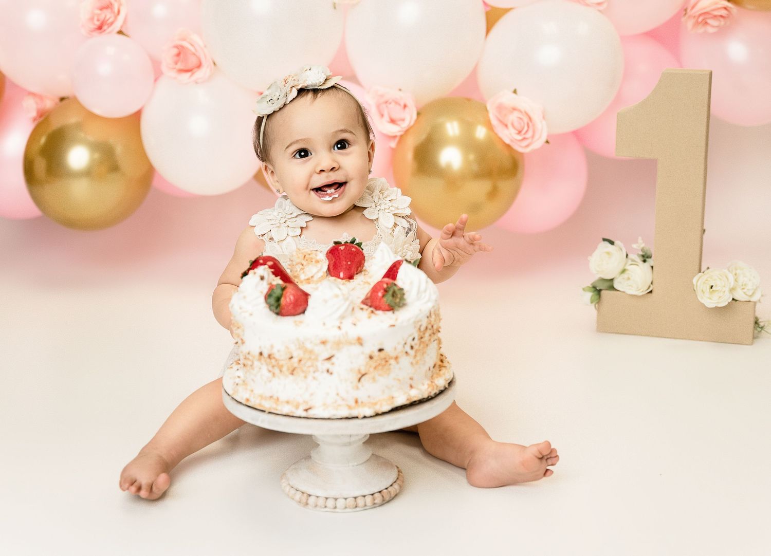 First birthday celebration setup with pink and gold balloons and a white cake on a pedestal.