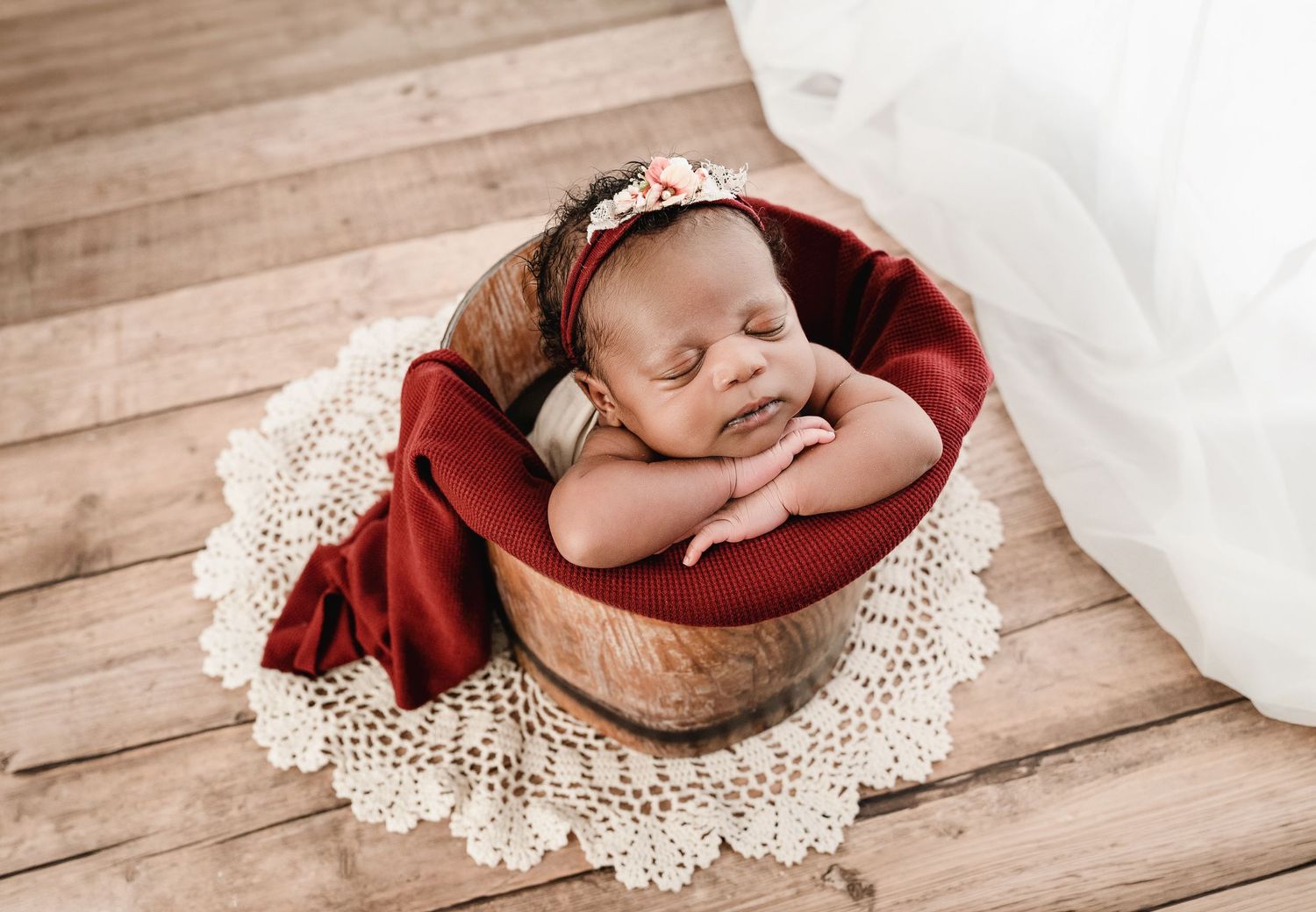 Newborn baby sleeping peacefully in a wooden bucket with red wrap and floral headband on lace doily.
