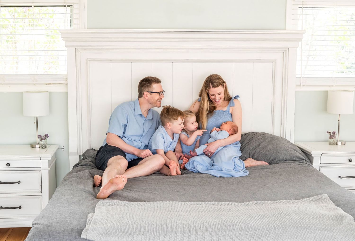 Family of four sitting on a bed, parents and two young children in coordinating blue outfits, in a bright white bedroom.