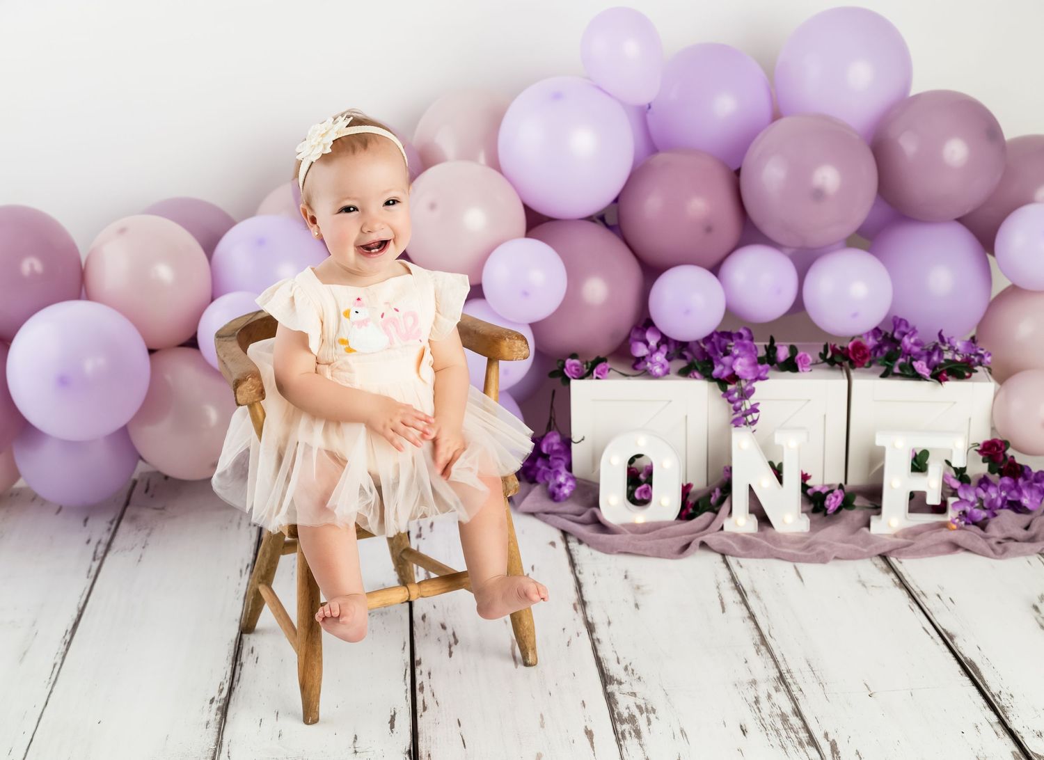 Baby girl in white tutu sits on chair surrounded by purple balloons and ONE letters at her first birthday cake smash.