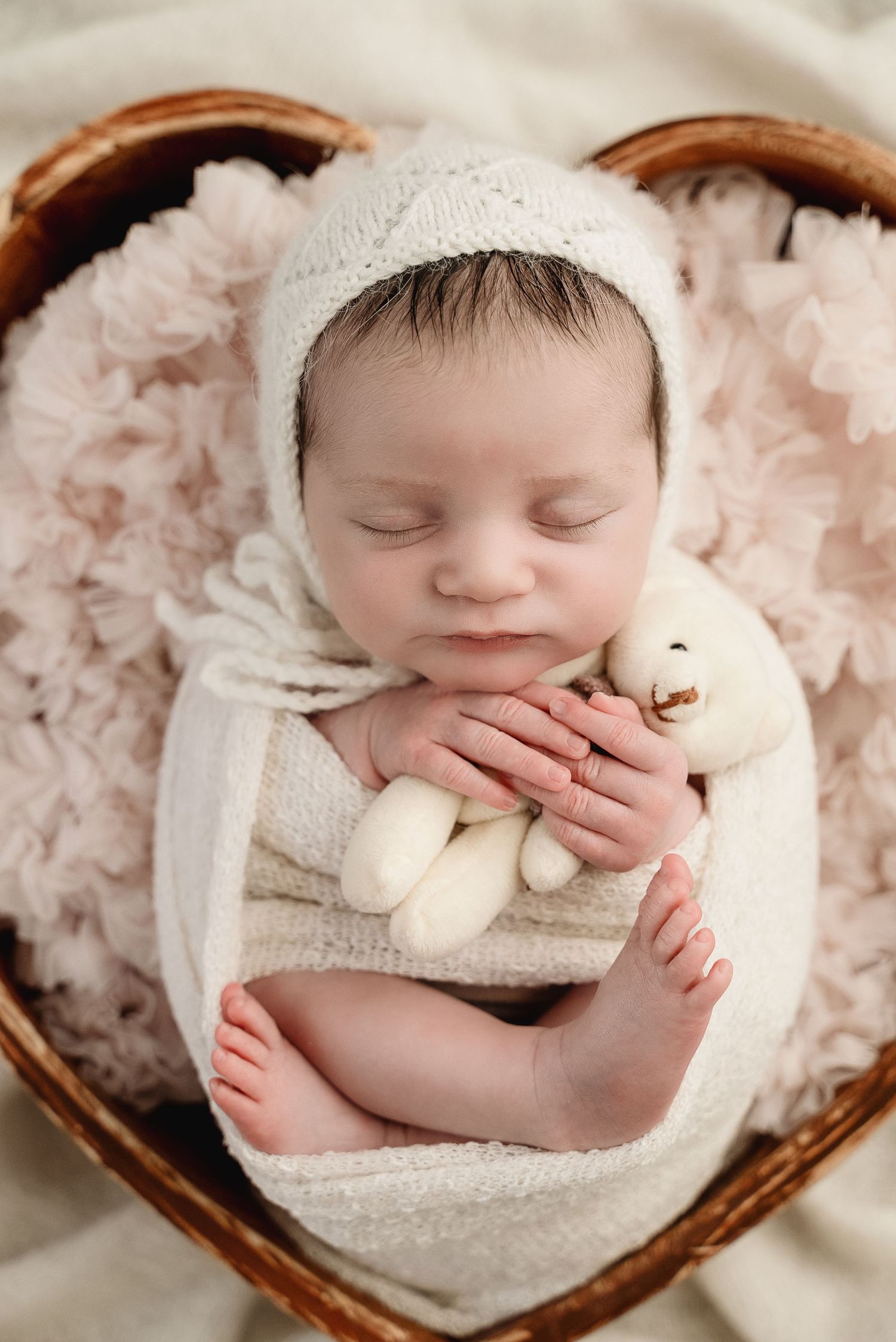 Sleeping newborn wrapped in soft cream fabric in wooden bowl with delicate staging.