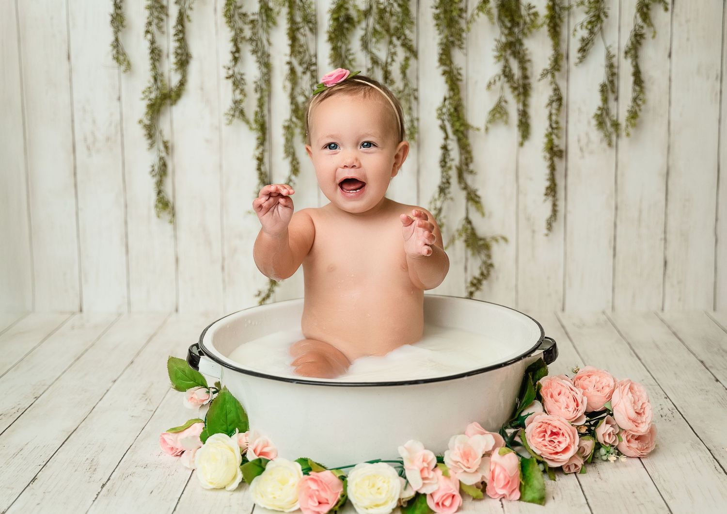 Baby sitting in vintage enamel bathtub surrounded by pink roses and greenery.