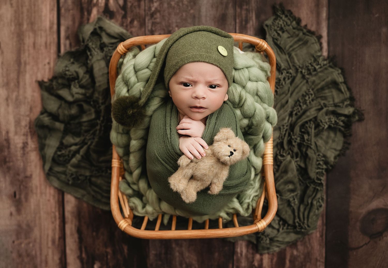 Baby in olive green outfit sleeping in wicker basket with plush teddy bear.