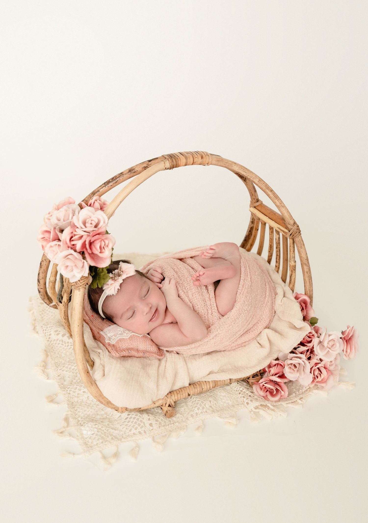 A sleeping newborn wrapped in pink fabric lies in a wooden basket adorned with delicate pink roses and lace details.