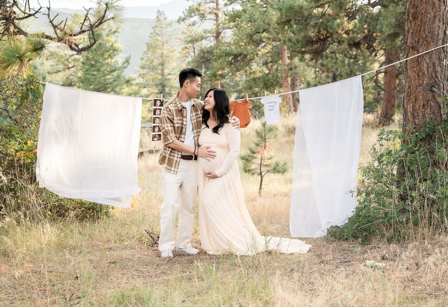 A couple walks through a sunlit forest with white sheets hanging between pine trees creating a dreamy outdoor scene.
