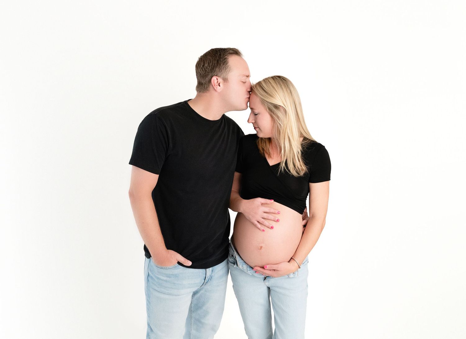 Couple in matching black shirts, man kissing pregnant woman's forehead while she cradles her baby bump.