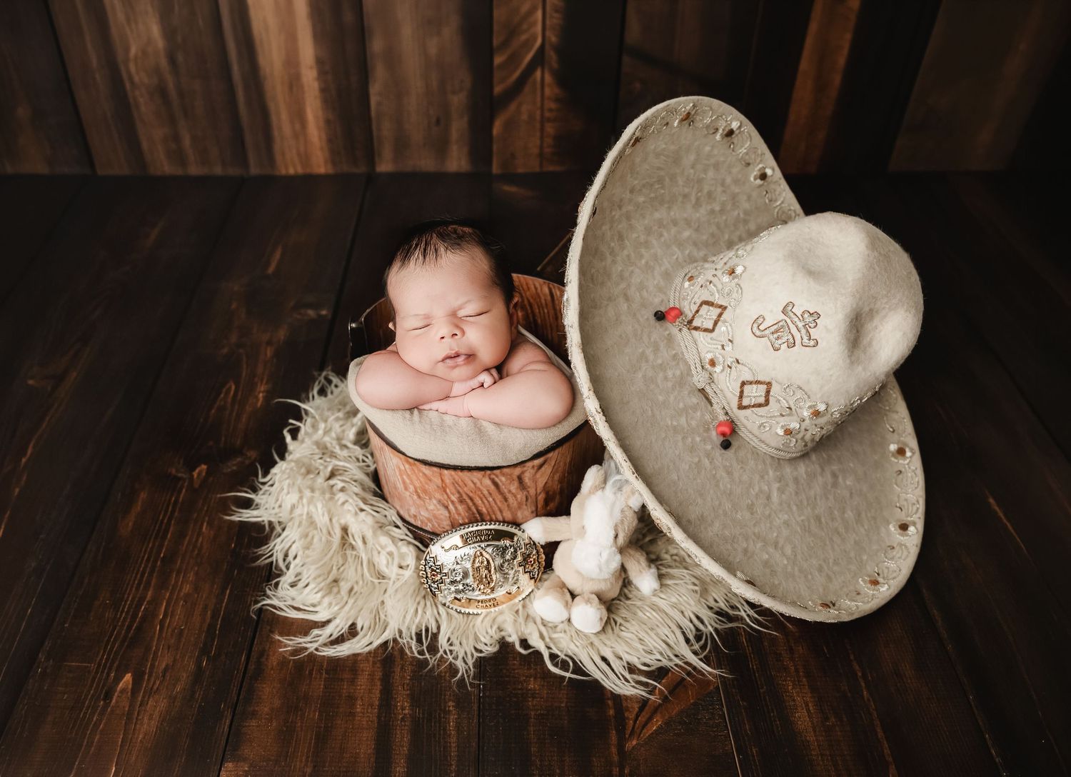 Newborn baby sleeping in a wooden bowl with a cowboy hat and toy bunny on a rustic dark wood background.
