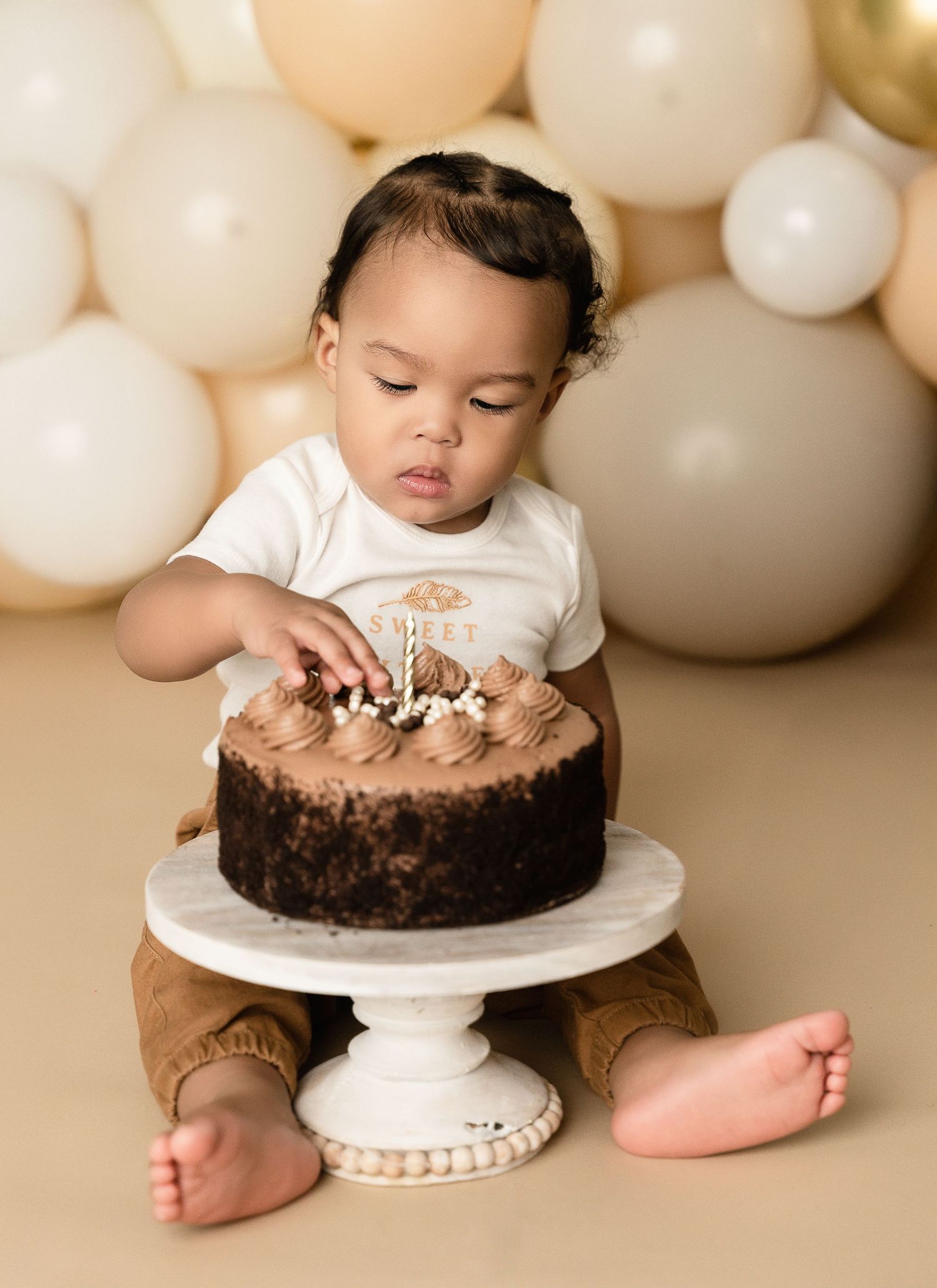 First birthday cake smash photo with chocolate cake on white pedestal against balloon backdrop.