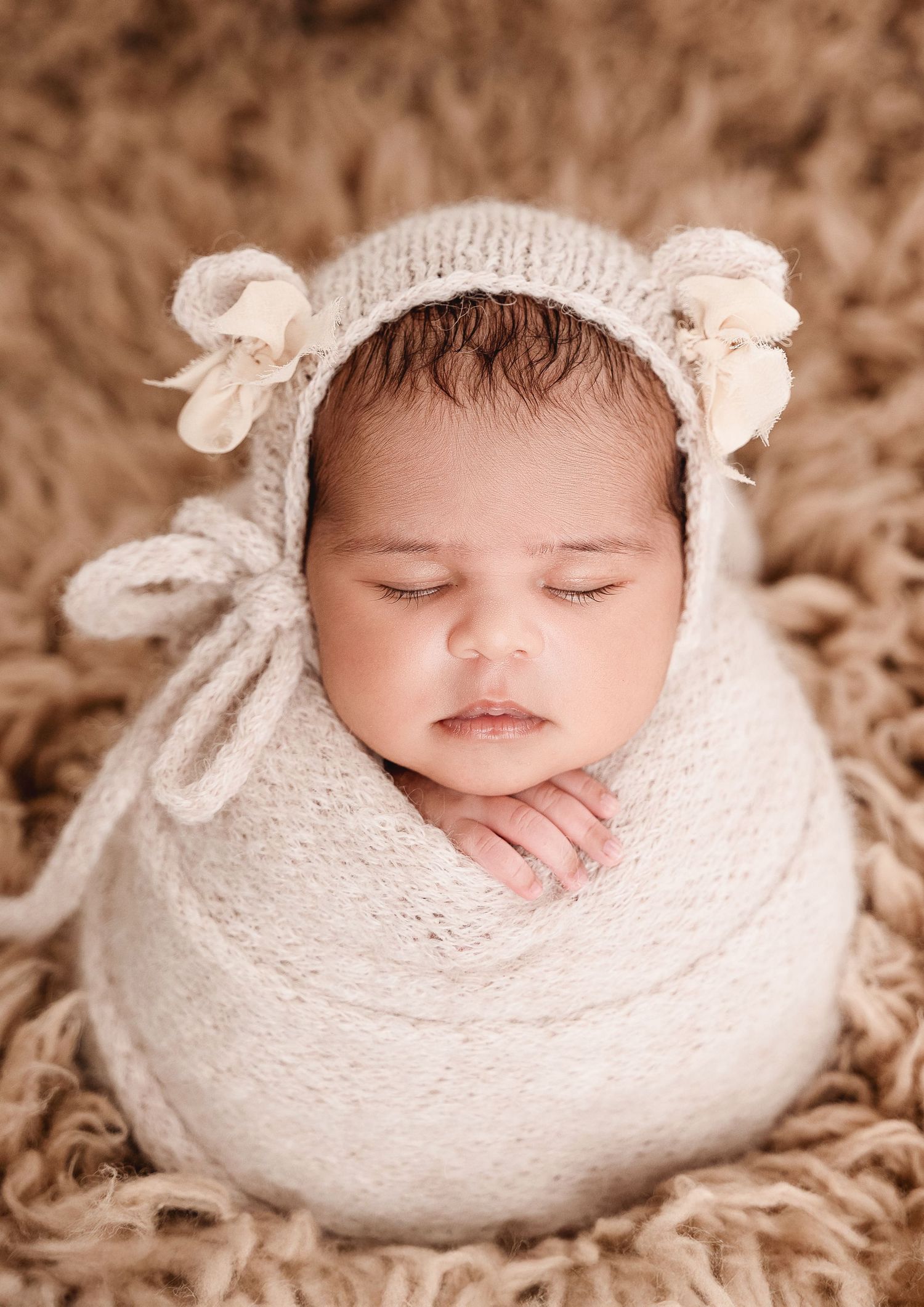 Sleeping newborn wrapped in soft white knit blanket with bear ear bonnet on brown textured background.