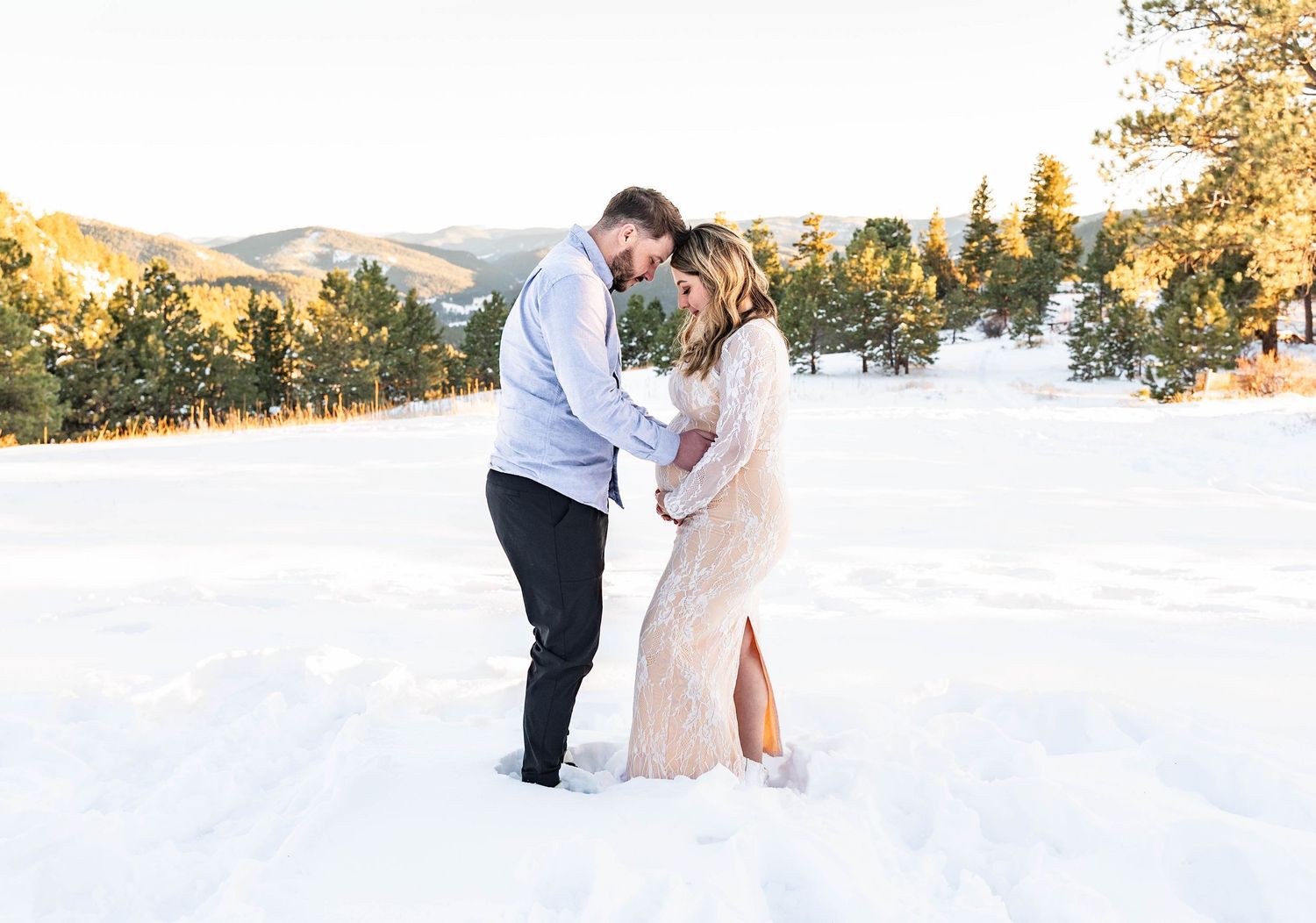 A couple embraces in the snow during sunset with mountains in the background.