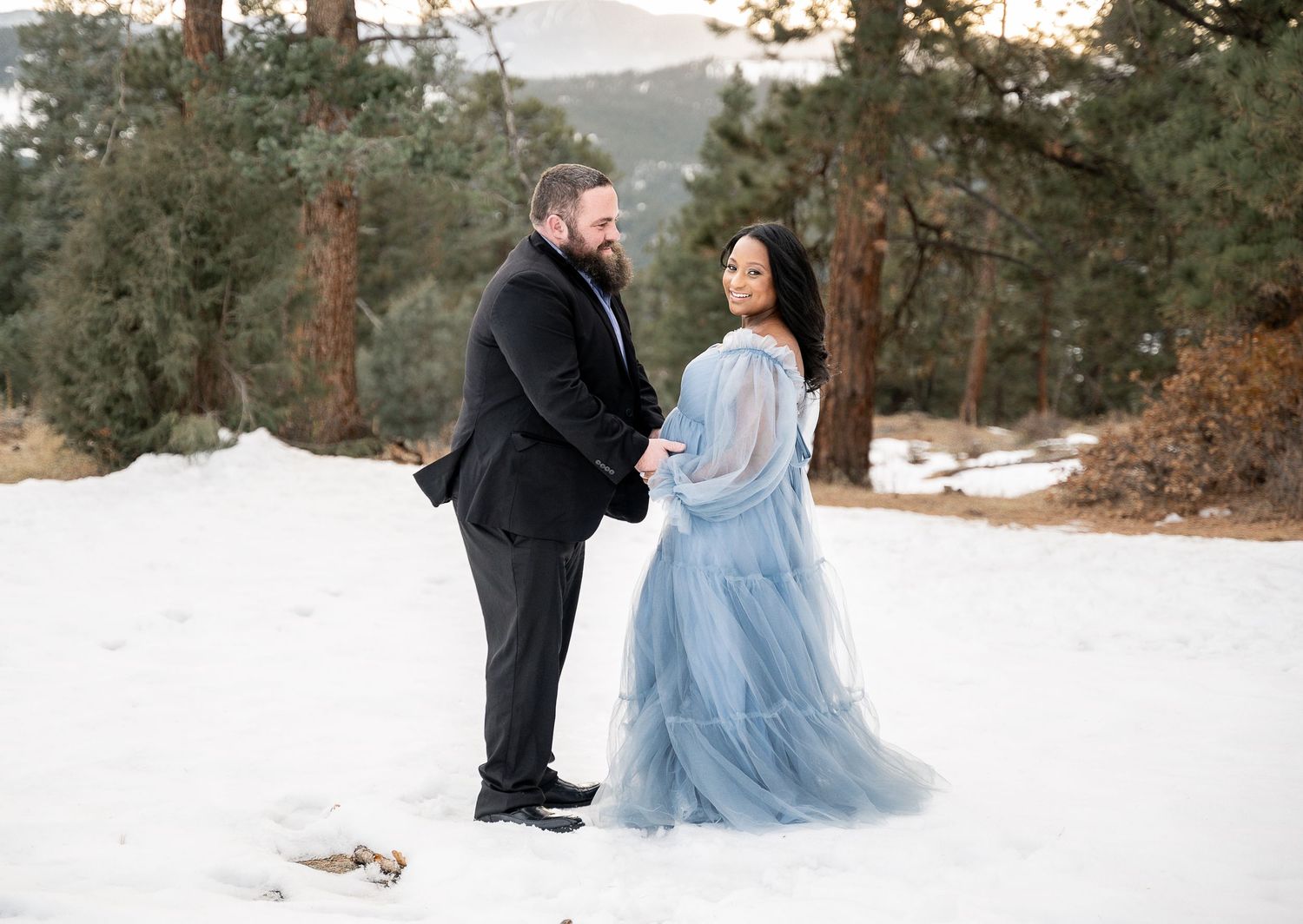Winter maternity photoshoot with couple in formal wear standing in snowy forest.