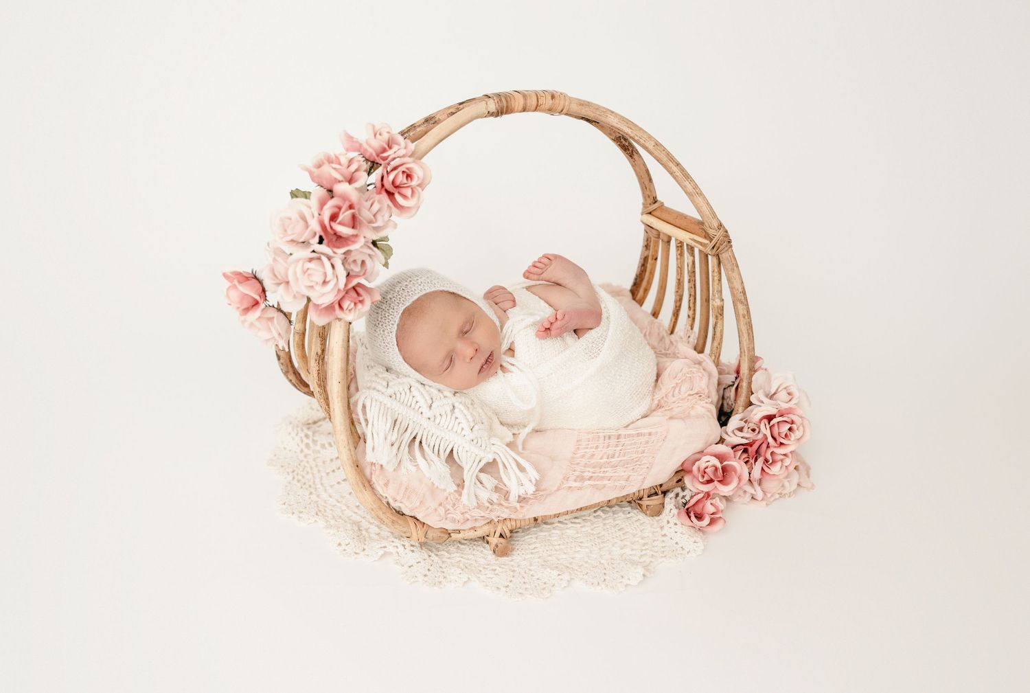 Delicate pink roses frame a soft white bassinet in a dreamy newborn photography setup.