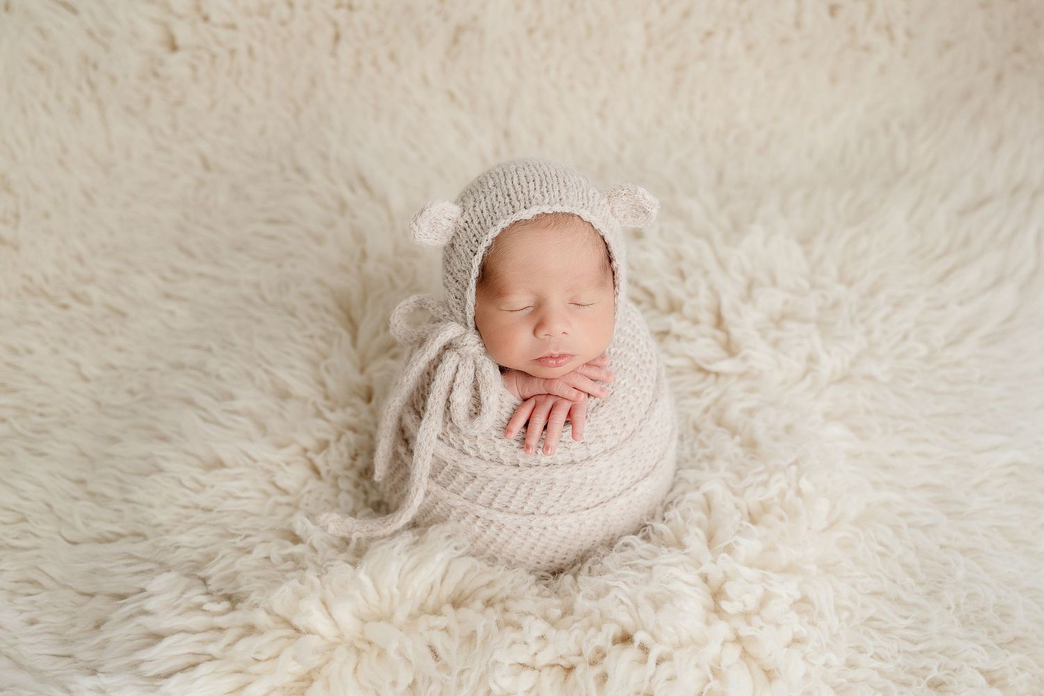 A sleeping newborn wrapped in a cream knit blanket lies on a soft white fur background wearing a lace bonnet.