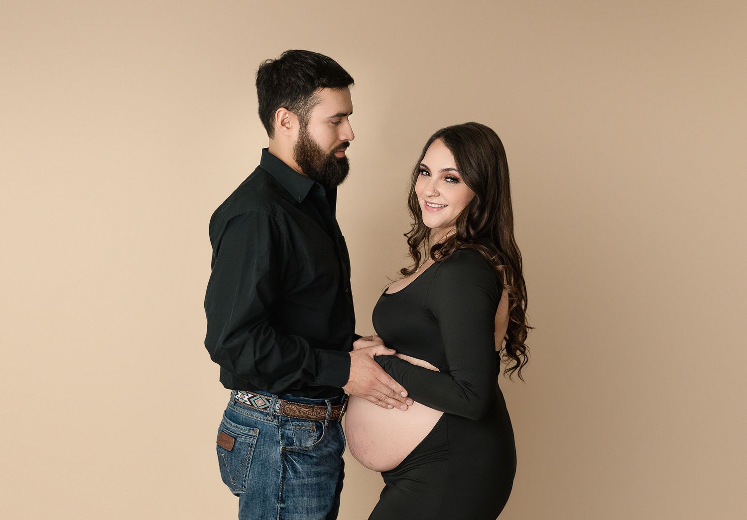 A couple stands together against a beige backdrop during a maternity photo session.