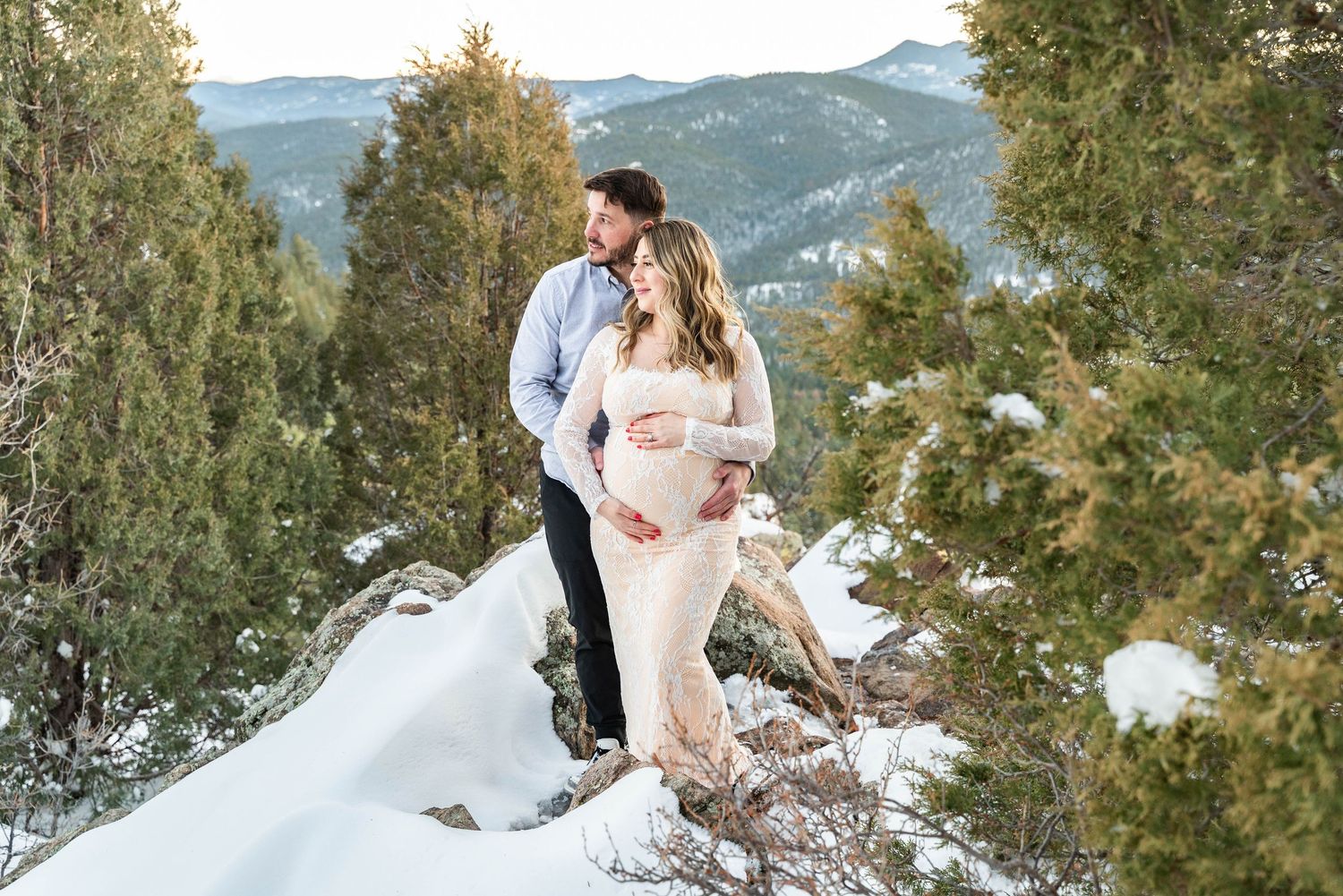 A couple in wedding attire poses in a snowy mountain landscape with evergreen trees in the background.