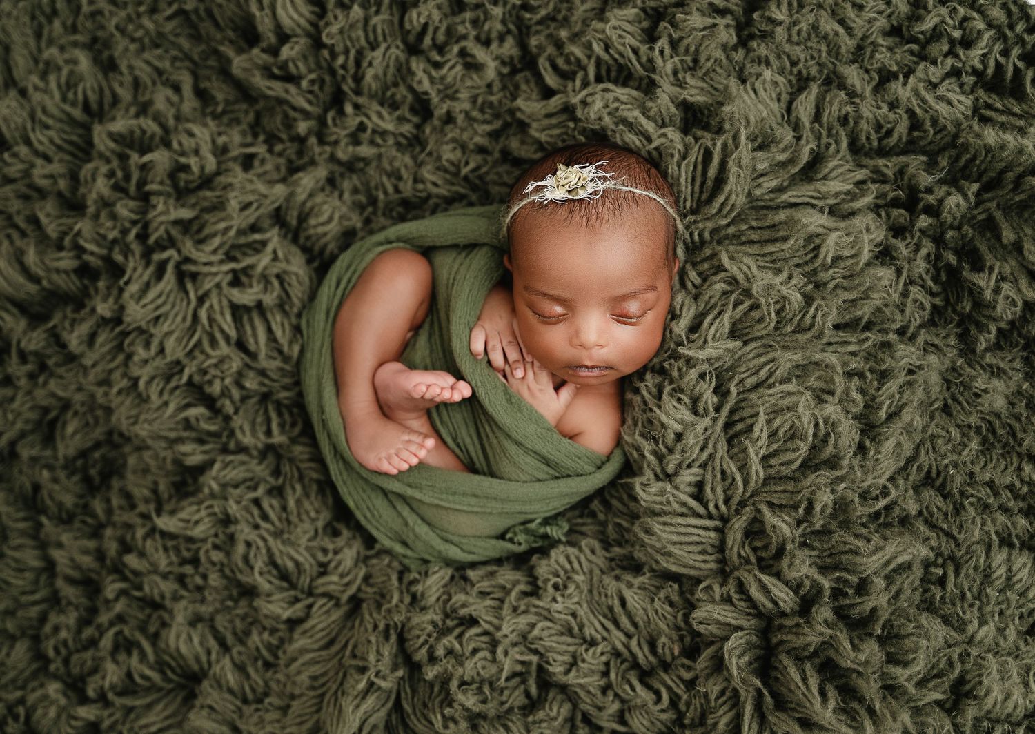 Overhead view of a sleeping infant wrapped in olive green fabric on textured carpet.