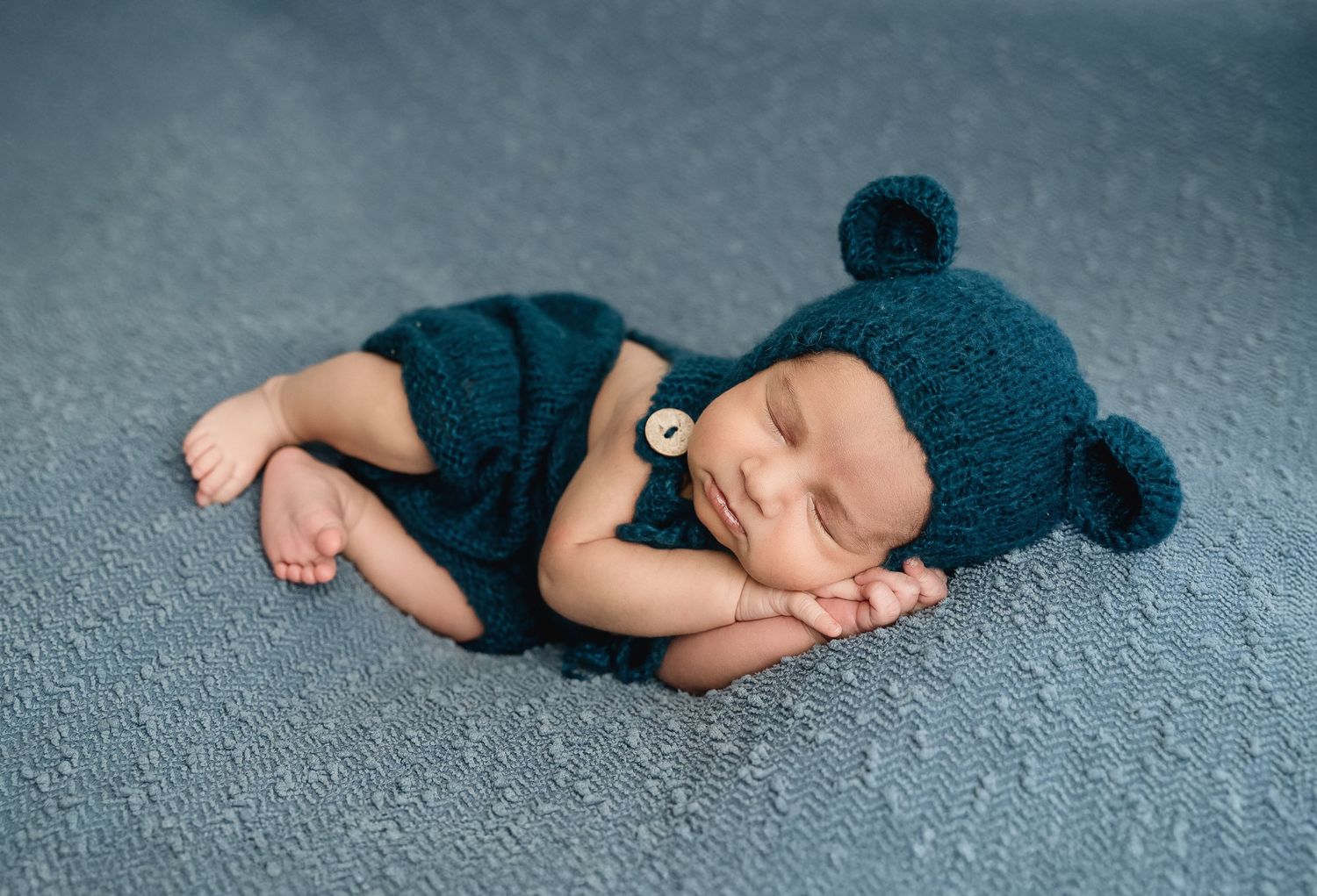 Sleeping baby wearing blue knit bear hat and shorts on blue textured backdrop.