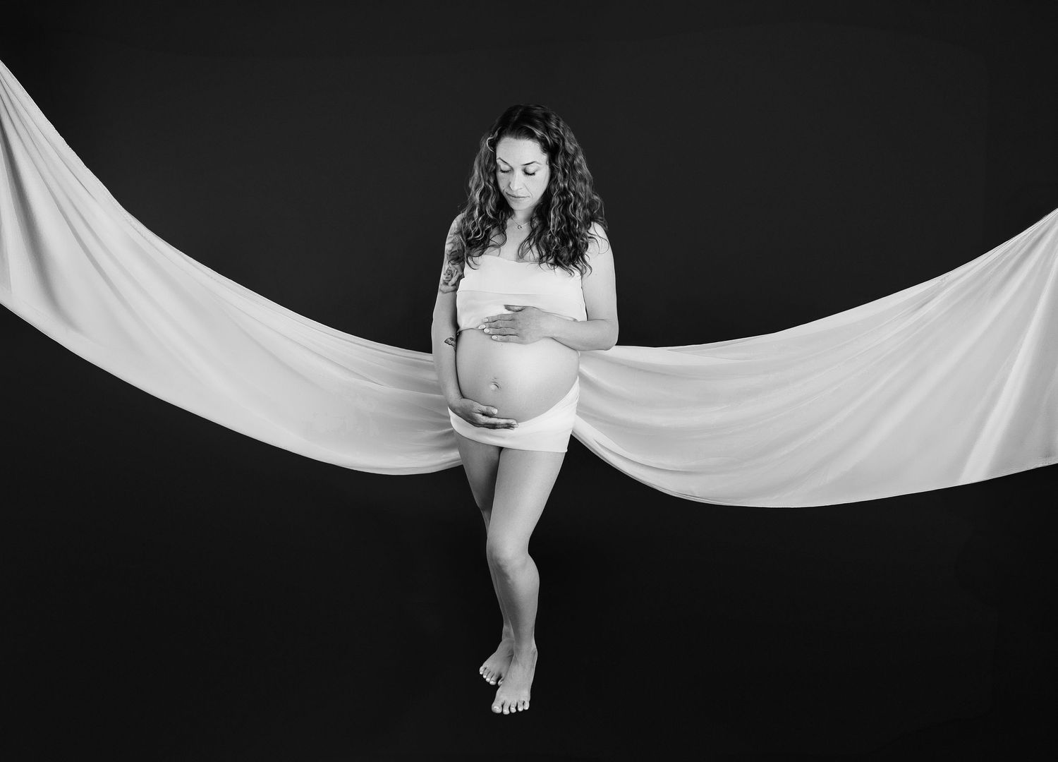 Black and white maternity photo of a pregnant woman draped in white fabric against a dark background.
