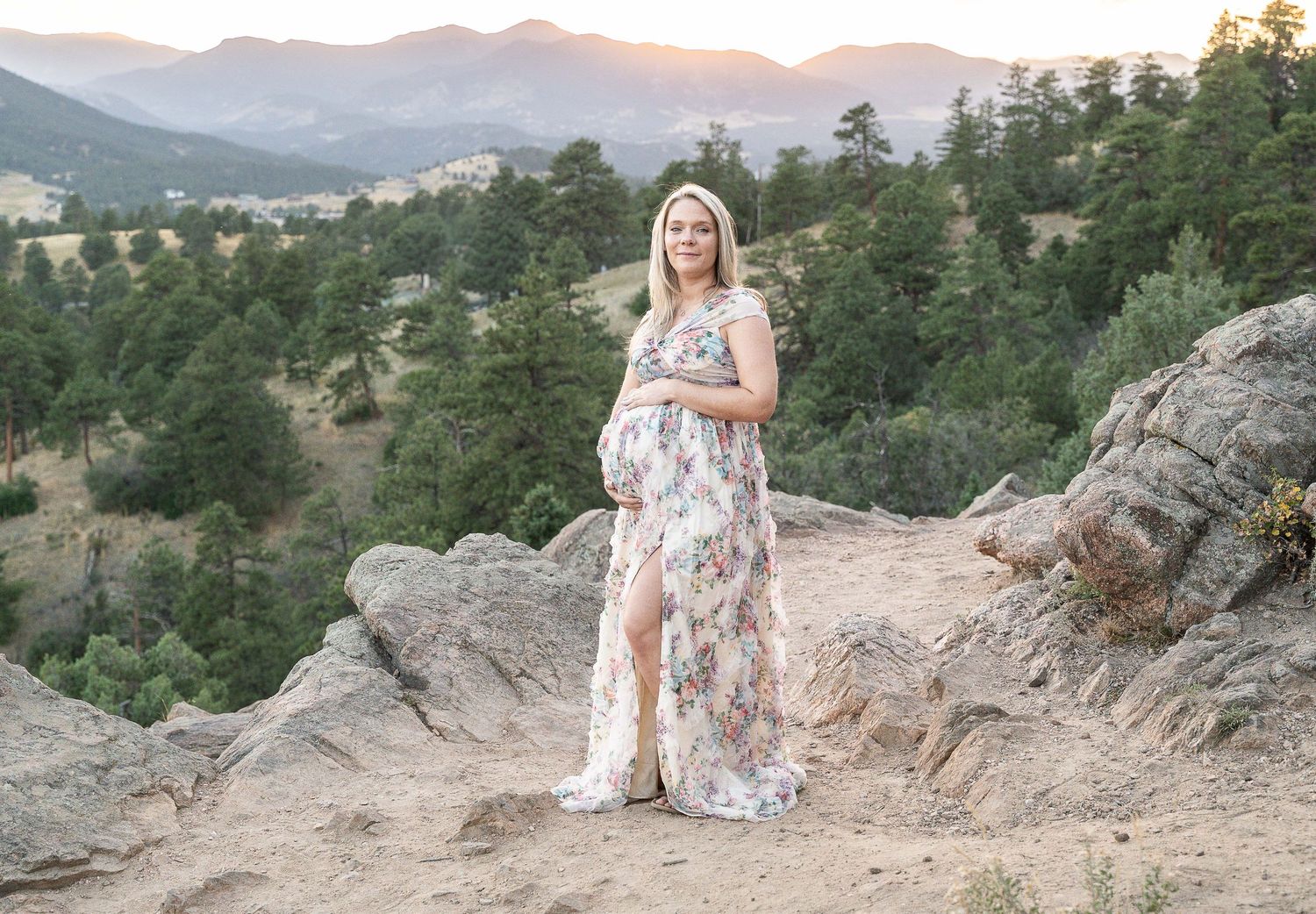 A person in a white floral dress poses on rocky mountain overlook at sunset with pine trees in the background.