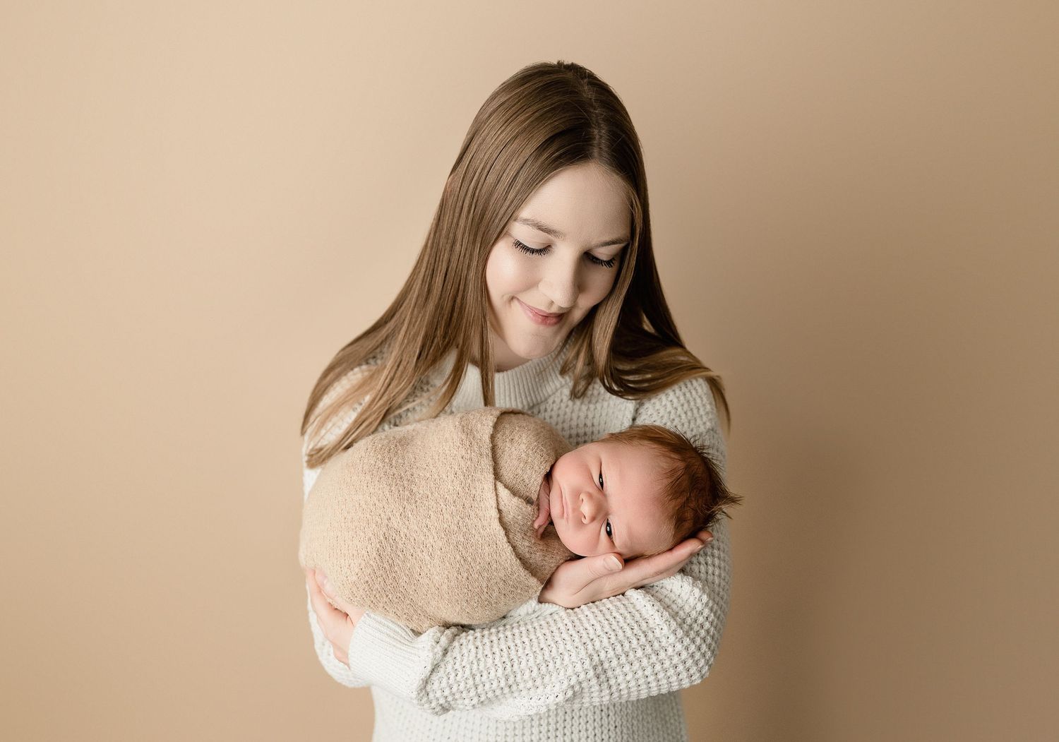Mother in beige sweater holding sleeping newborn against neutral backdrop.