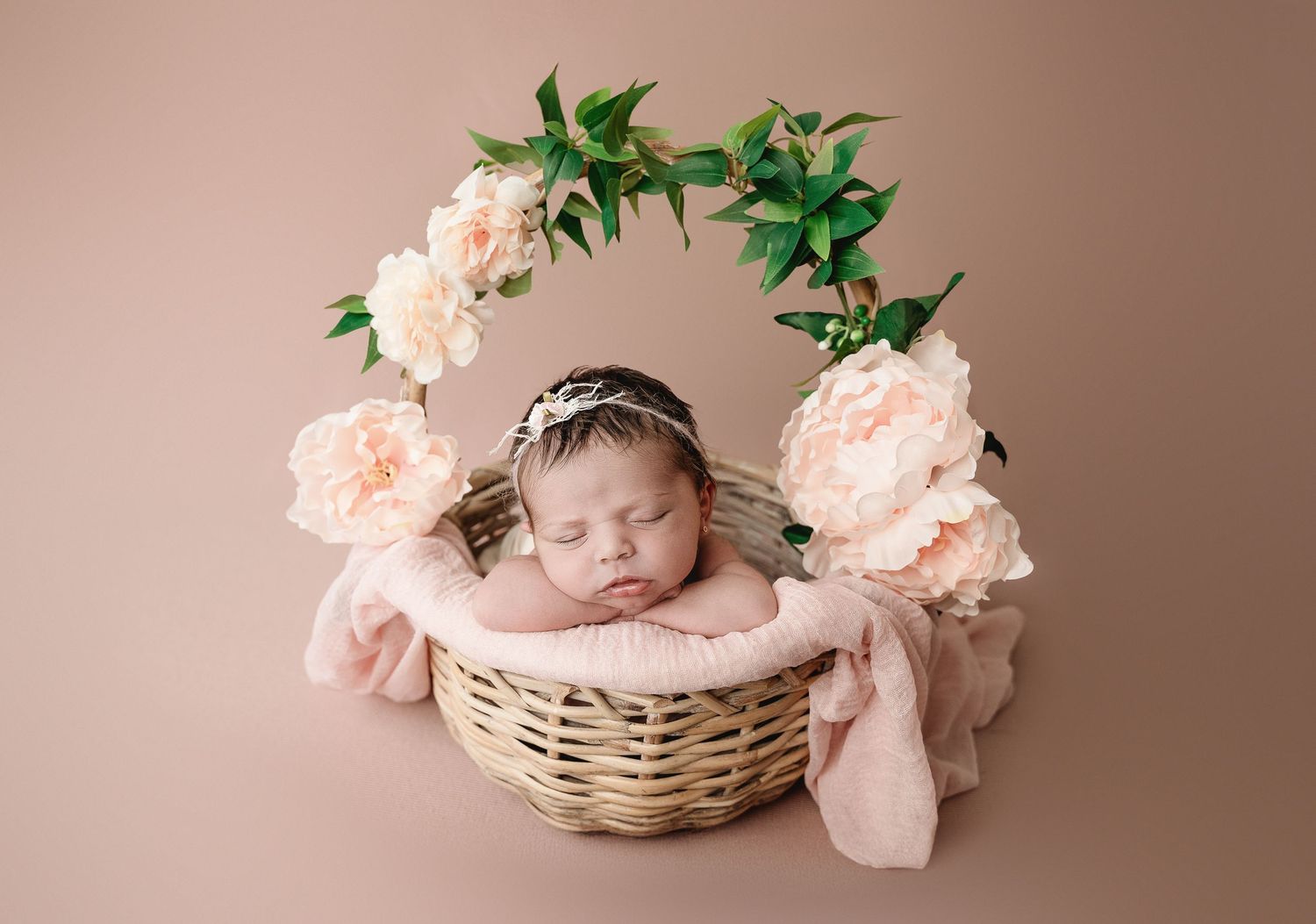 Sleeping newborn nestled in wicker basket surrounded by soft pink peonies and greenery on beige backdrop.