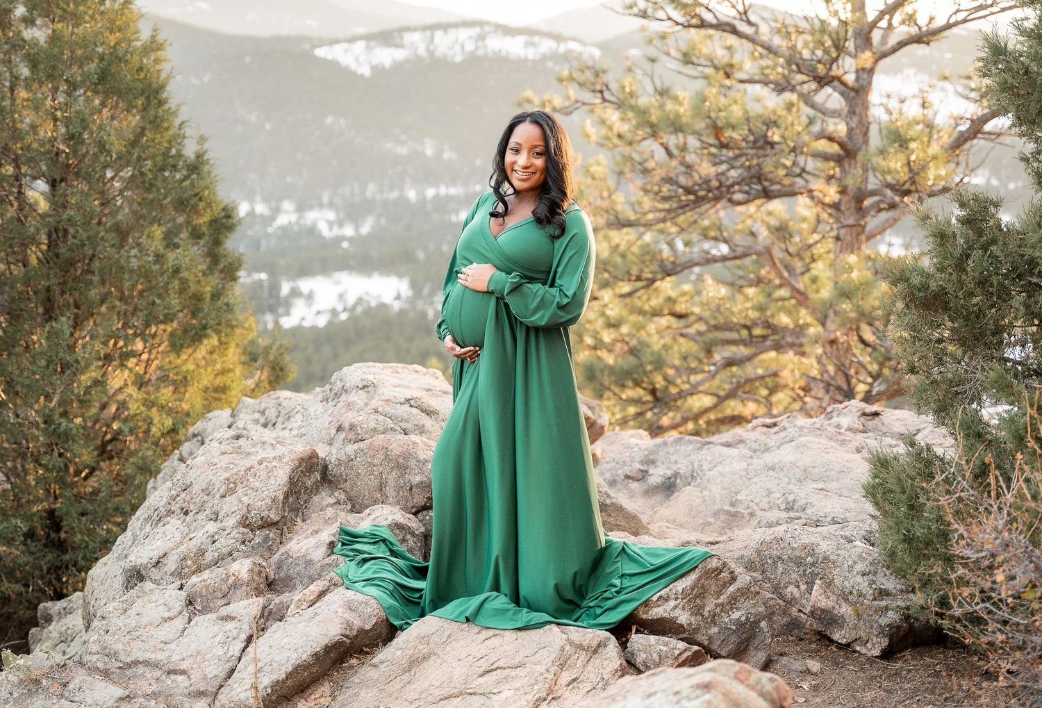 Maternity photo of emerald green dress on mountain cliff with scenic backdrop.