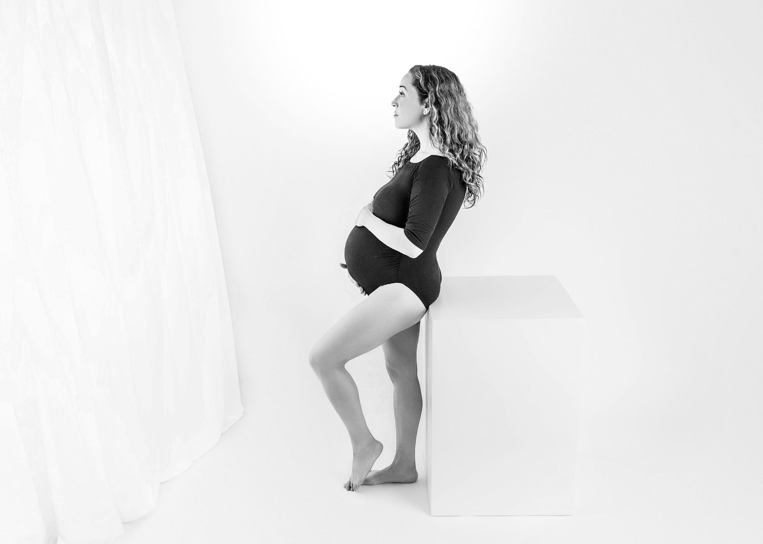 Black and white maternity photo of a pregnant woman in a bodysuit leaning against a white cube in a bright studio.