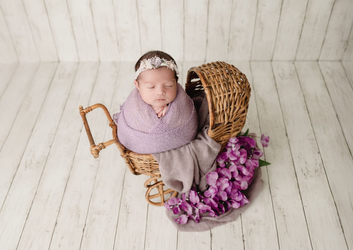 A newborn baby in a purple wrap lies next to wicker baskets filled with purple flowers on a white wooden floor.