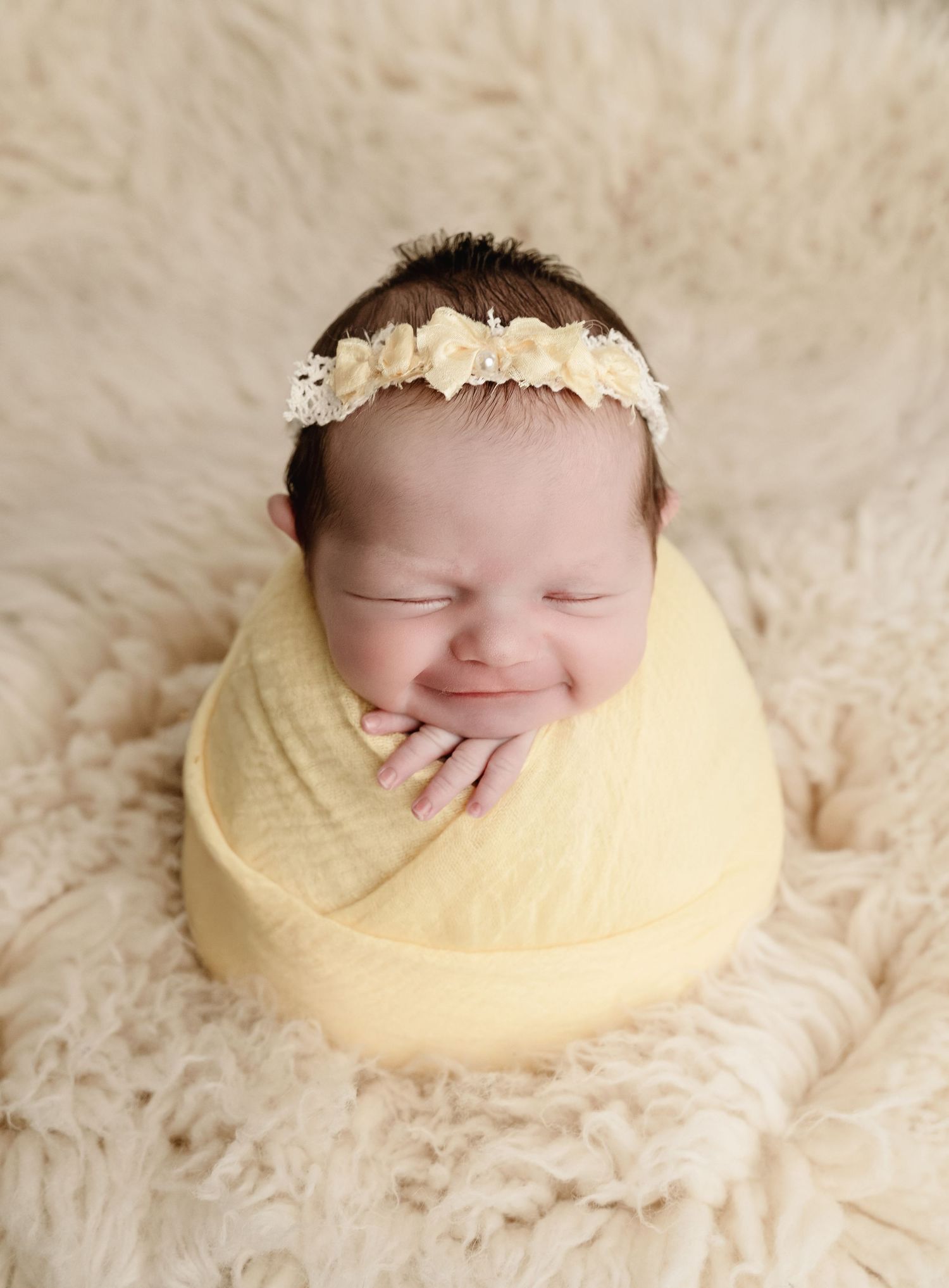 Sleeping newborn baby wrapped in yellow swaddle with floral headband on fluffy cream background.