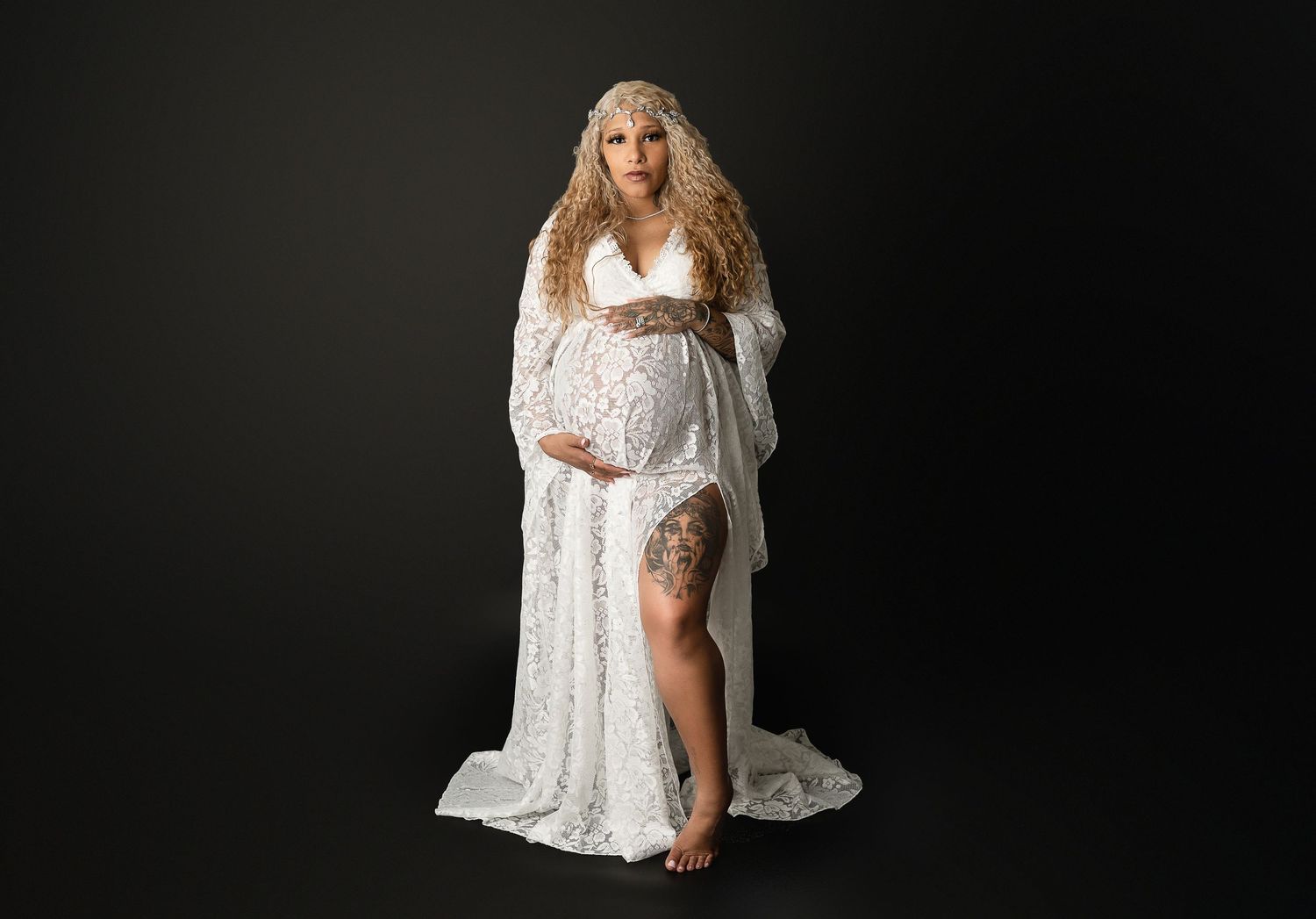Dramatic maternity portrait in white lace gown with leg slit against dark background.