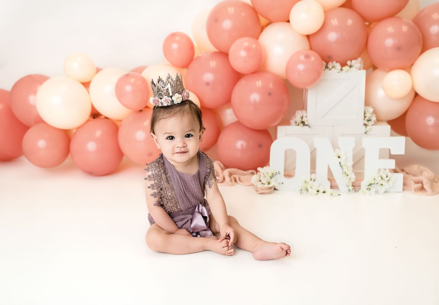 A first birthday celebration setup with pink balloons, white decorative letters, and an adorable baby in purple.Cake smash session in Denver