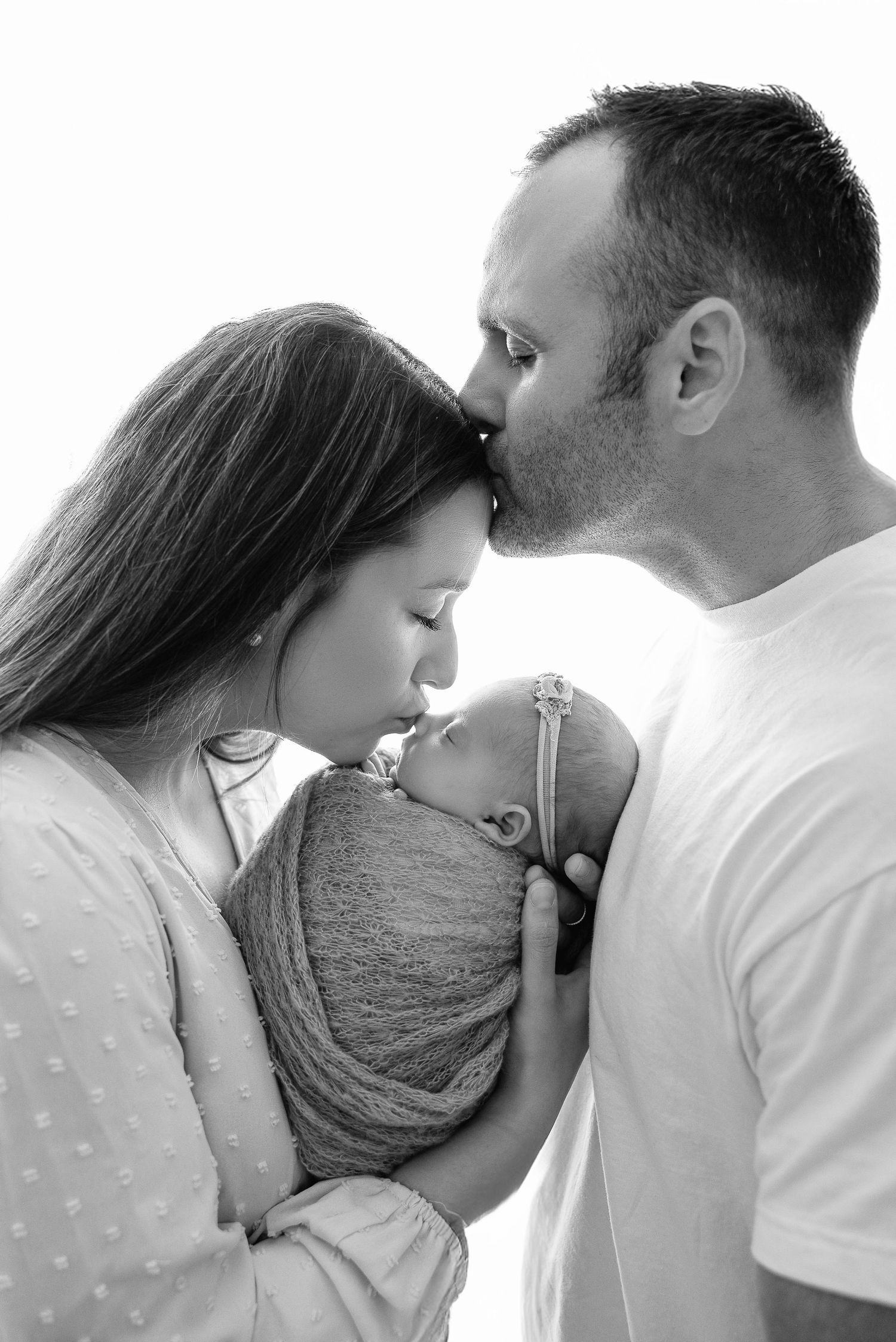 Black and white portrait of parents sharing tender moment with newborn baby wrapped in soft fabric.