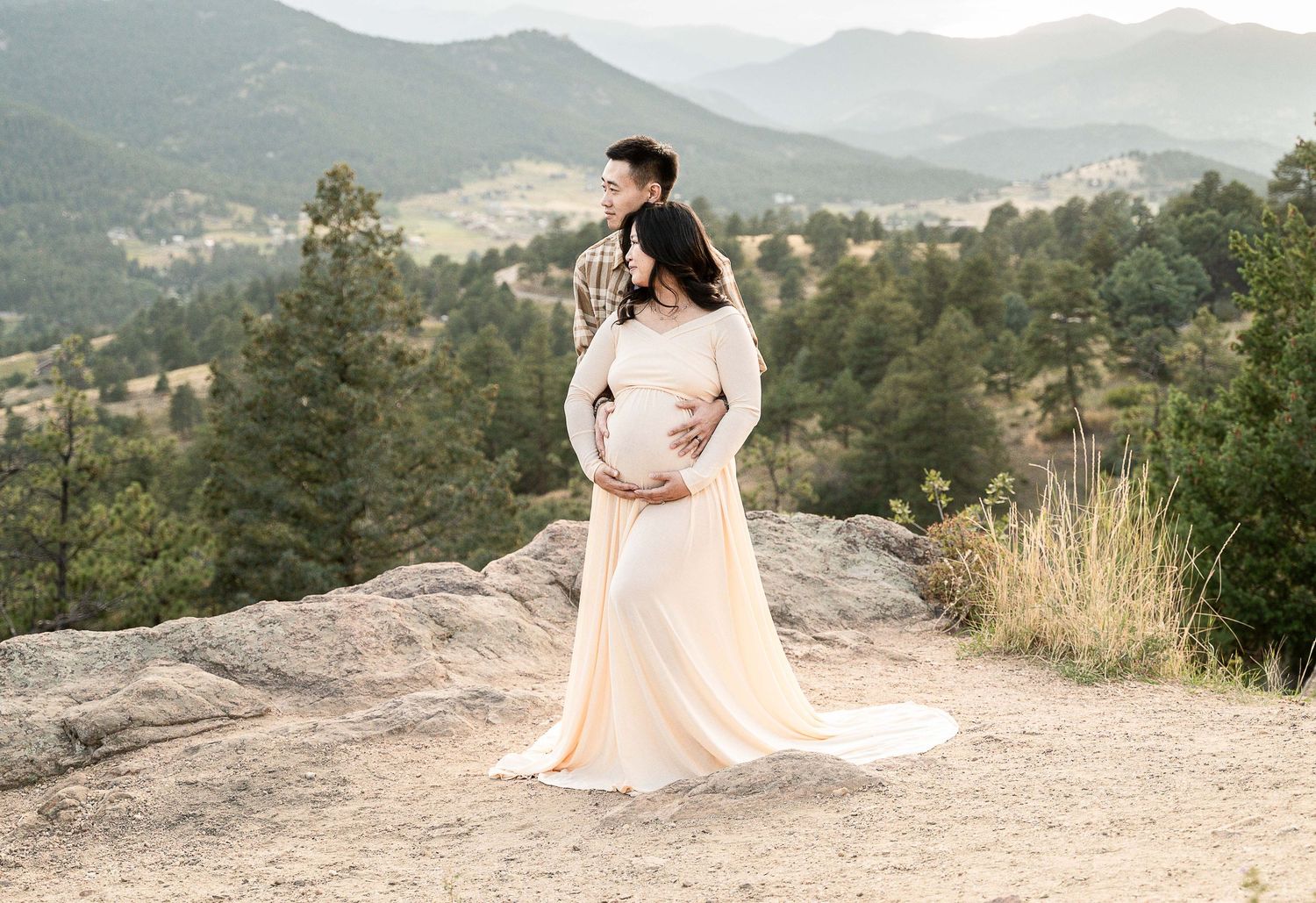 A romantic couple embraces on a rocky mountain overlook with scenic mountain vistas in the background at sunset.