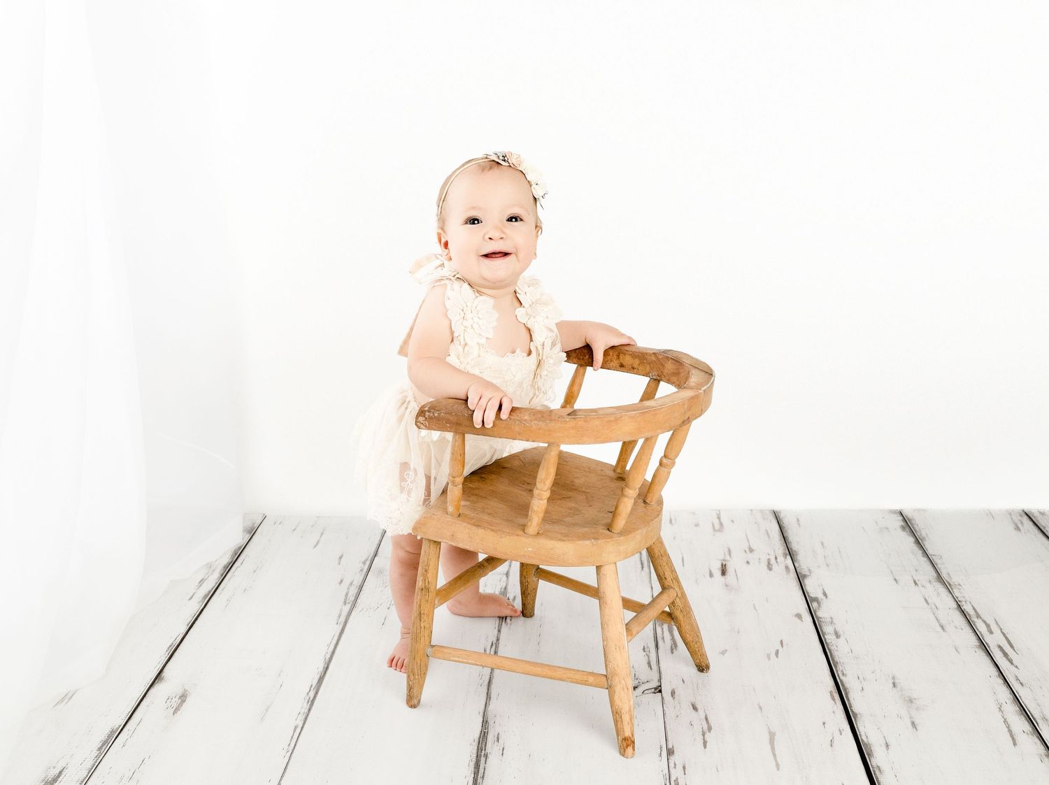Smiling baby girl in pink romper standing beside a wooden chair on white rustic floors in a bright studio.